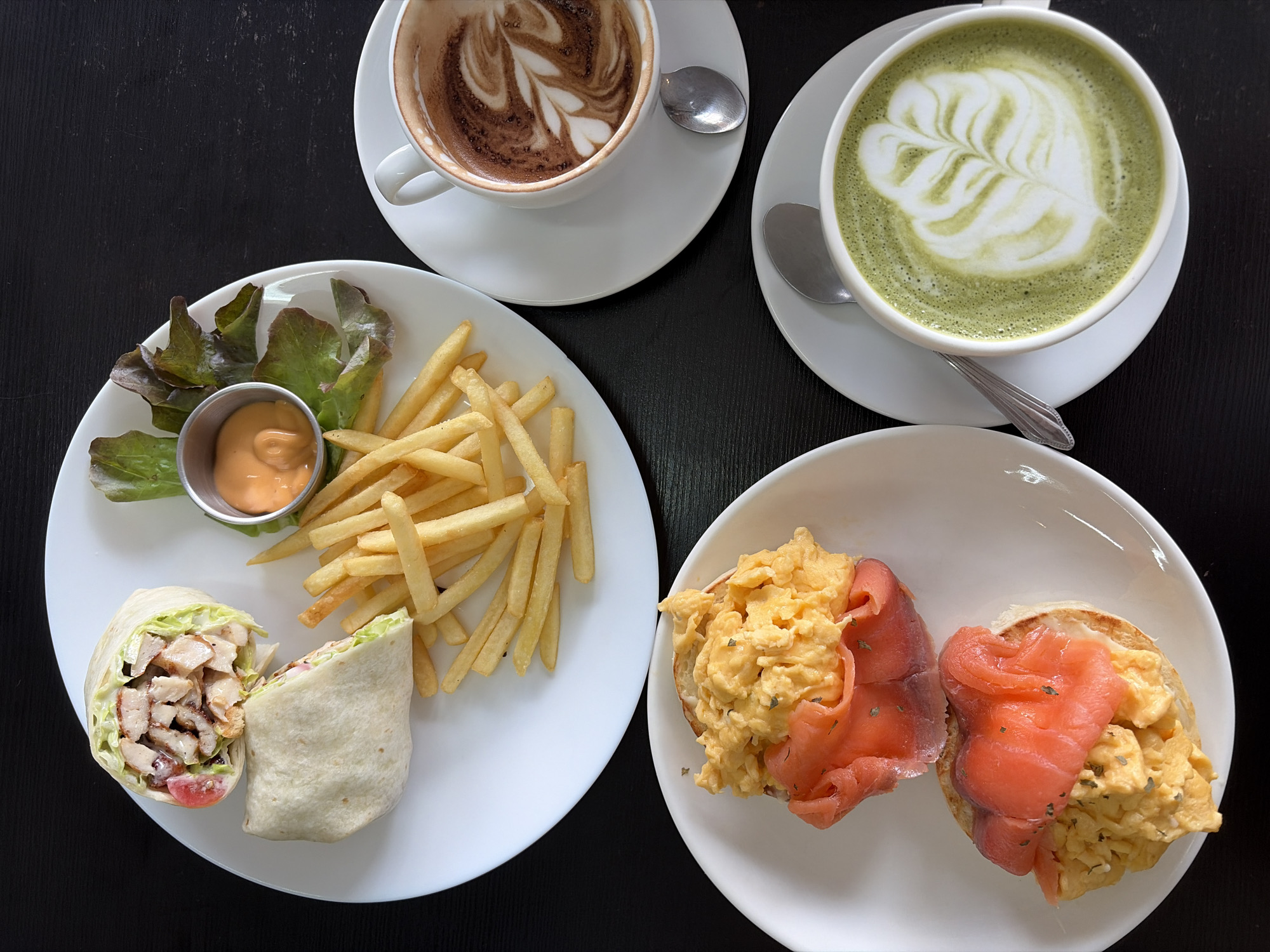 A high-angle, full shot of a table with food and drinks is captured on a dark wooden surface, possibly in Bangkok, Thailand. In the upper left, a white plate holds a chicken wrap, cut in half, with french fries and a small cup of dipping sauce. Adjacent to this, on the right, a white cup with a saucer contains a latte with intricate foam art, and a spoon is visible. Below the latte, another white cup with a saucer holds a matcha latte, also with foam art, and a spoon. In the lower left, a white plate features two halves of an English muffin topped with scrambled eggs and slices of smoked salmon. The scene suggests a breakfast or brunch setting.