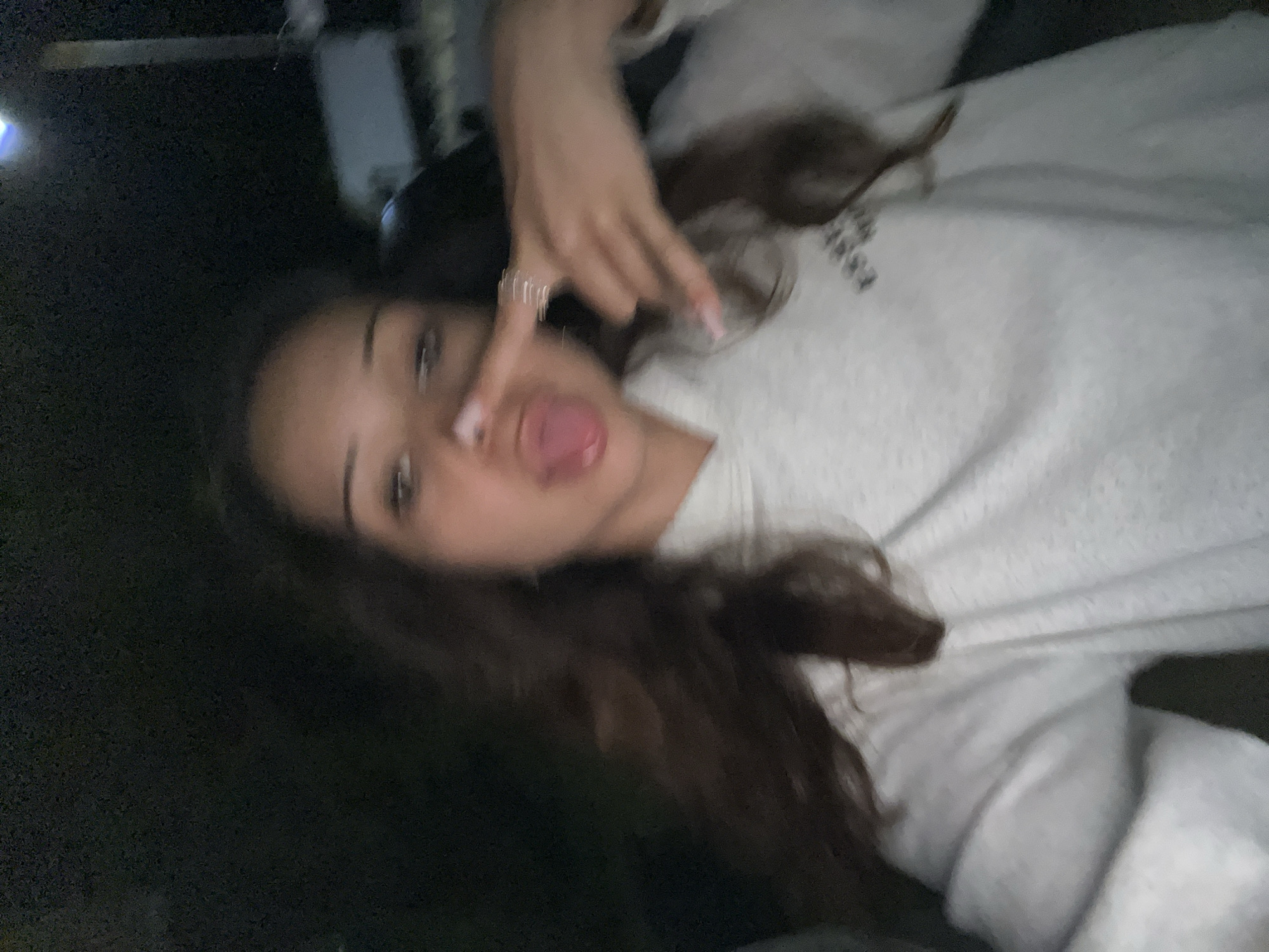 The image depicts a close-up, low-light selfie of a single individual, appearing to be a young female, in an outdoor or semi-outdoor setting at night or in very dim light, within the city of Unknown, Unknown. The individual has dark, wavy hair and is wearing a light-colored hooded sweatshirt with a partially visible dark text or logo on the chest. She is making two distinct gestures: her tongue is extended from her mouth, and her right hand (viewer's left) is raised in a "devil horns" or "rock and roll" gesture, displaying pink manicured nails and at least one ring. Her left hand (viewer's right) has its index finger touching her nose. The background is dark and heavily blurred, revealing indistinct elements such as a vertical structure, a faint blue light source in the upper right, and a light-toned, unidentifiable object, possibly a vehicle or building, in the middle-right distance. The overall image exhibits characteristics of a photo taken with limited illumination, resulting in some grain and blur.