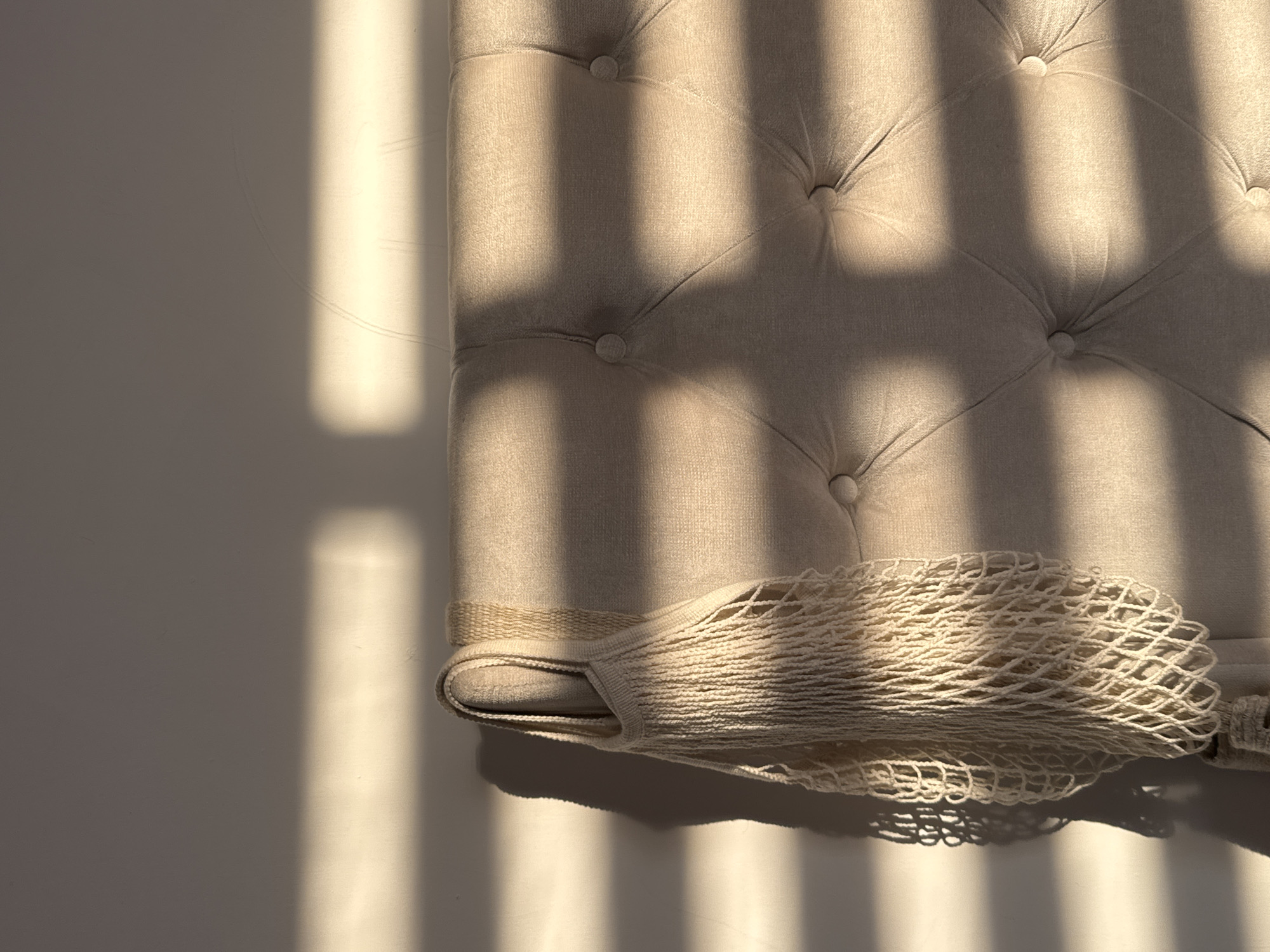The image presents a close-up view of a tufted, beige headboard with a cream-colored mesh bag draped over the lower edge. Light streams in from a window, casting distinct, parallel shadows across the headboard and the adjacent wall, creating a play of light and shadow. The tufting pattern consists of symmetrically arranged buttons embedded in the fabric. The mesh bag has a woven handle. The wall beside the headboard is white, providing a stark contrast with the shadows. The scene does not suggest a location in San Bernardino Tlaxcalancingo, Mexico as it is a close up of an interior setting that could be anywhere.
