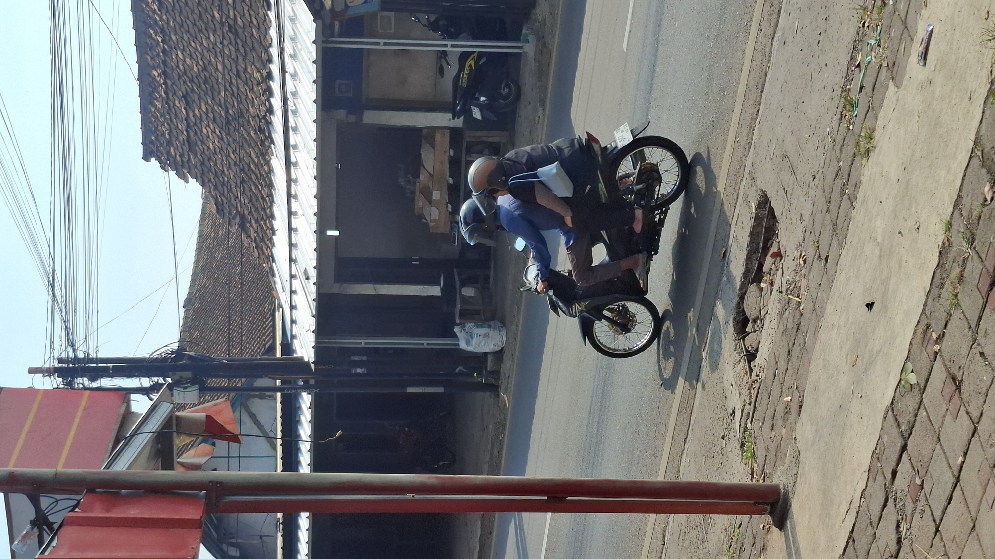 In Cilaku, Indonesia, on a sunny day, two individuals ride a motorcycle on an asphalt road. The rider, wearing a blue shirt and a dark helmet, operates the vehicle. The passenger, seated behind the rider, wears a dark shirt, a brown and black helmet, and carries a light blue bag on their right shoulder. The motorcycle is dark colored and has spoked wheels. A damaged section of the road surface, with exposed rebar and debris, is visible on the right side of the road. In the background, several buildings with tiled roofs line the street, some with metal awnings. Power lines crisscross the sky, supported by wooden utility poles. A red pole with yellow stripes stands prominently in the foreground on the left.