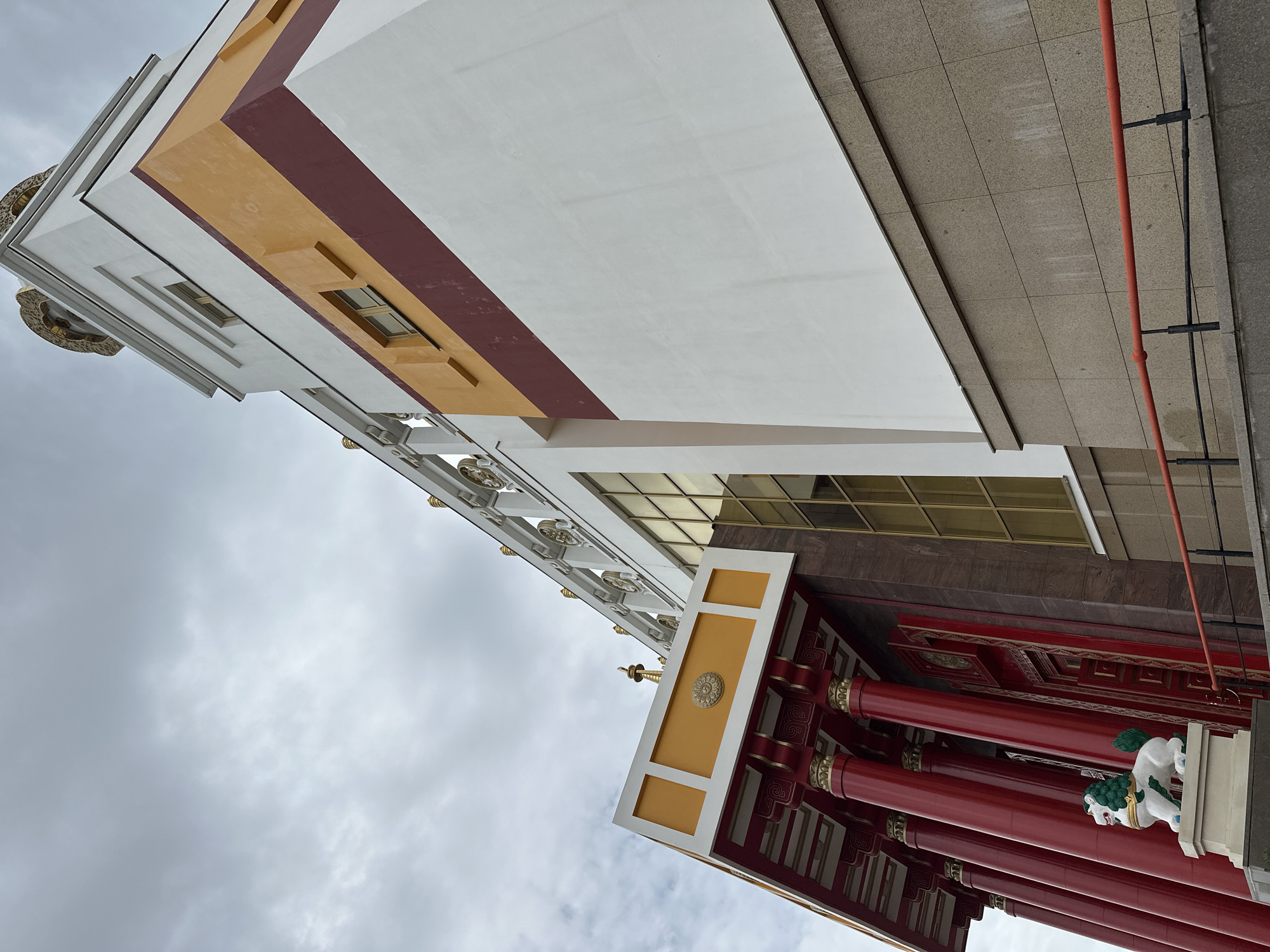 A low-angle, exterior shot captures a portion of the Golden Abode of the Buddha Kalachakra, located in Elista, Russia. The building features a striking architectural style with vibrant red columns supporting an ornate yellow and white overhang. The main facade of the structure is a stark white, accented by horizontal bands of maroon and ochre. Windows with golden frames punctuate the upper levels, and a decorative railing with intricate golden details runs along the roofline. In the foreground, a white lion statue with a green mane stands guard on a pedestal. The sky above is overcast with a diffuse gray light.