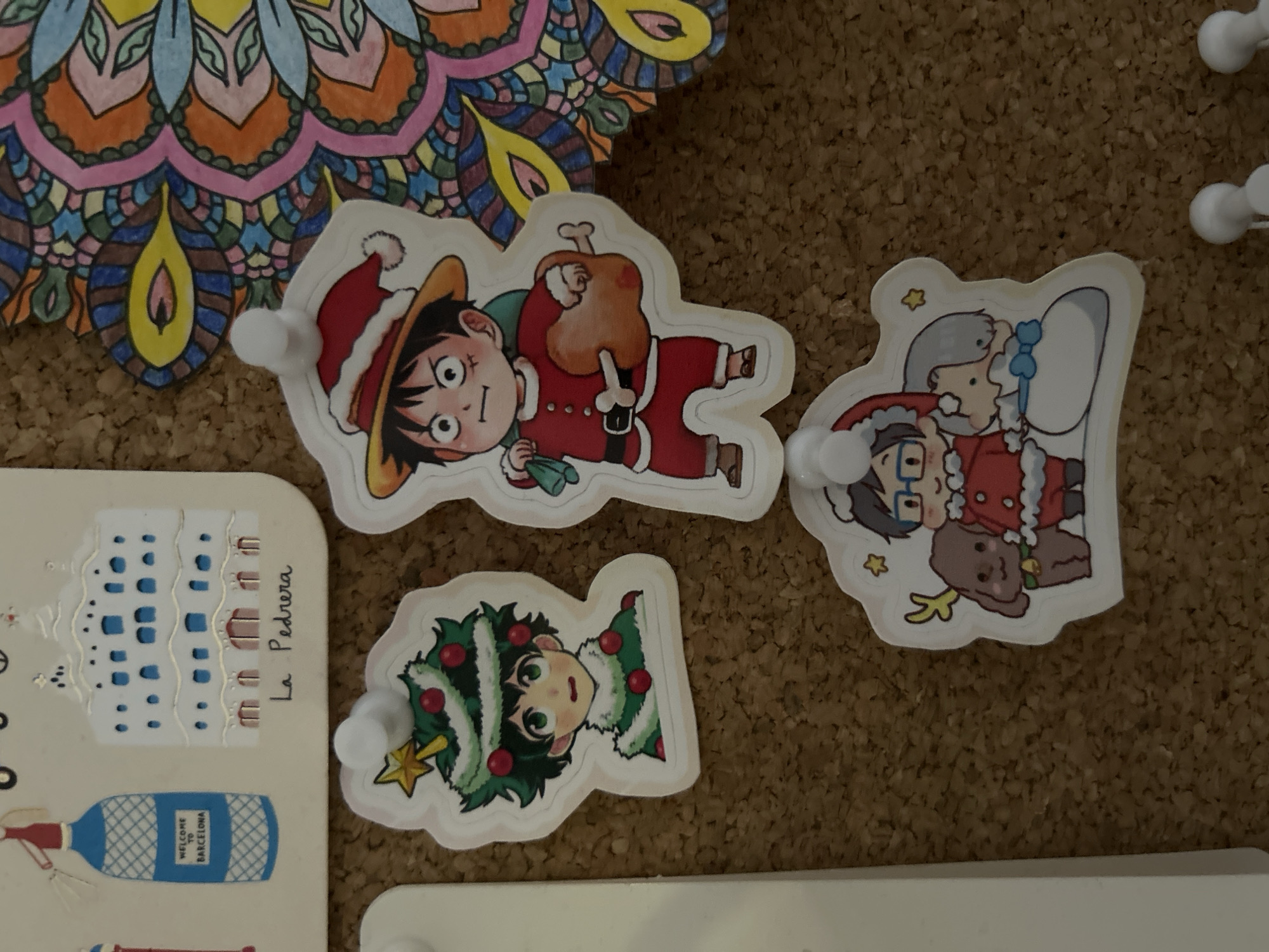 The image shows a corkboard with several items pinned to it. There is a partially visible colorful mandala drawing in the top left corner. Below the mandala is a postcard with a depiction of La Pedrera, a building in Barcelona, Spain, and a bottle labeled "WELCOME TO BARCELONA." Three anime-style character stickers, each depicting a different character dressed in Christmas-themed attire, are pinned to the board with white push pins. The background is a brown corkboard. The image provides no information about Câmara de Lobos, Portugal.