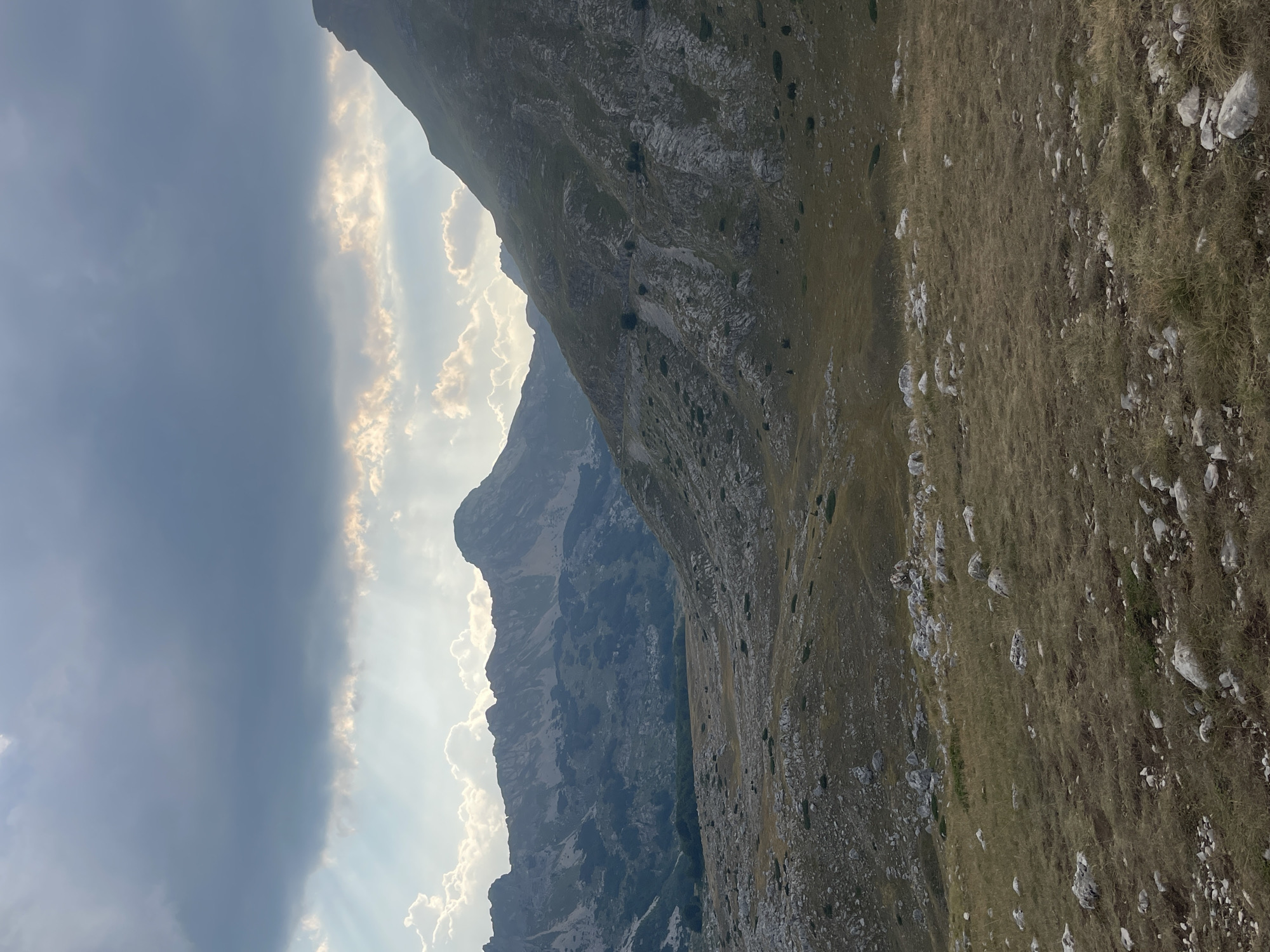 The image depicts a mountainous landscape under a dramatic sky. In the foreground, a grassy, rocky terrain slopes upwards. Scattered rocks and sparse vegetation characterize this area.
In the midground, the terrain transitions into a valley. The valley floor appears covered in grass and dotted with trees, some of which are dark green. The slopes of the mountains rise steeply on either side of the valley. The mountain on the right is covered in grass and rock, with visible ridges and gullies. The mountain range in the distance is rugged and appears to be made of rock, with visible peaks and sharp contours.
The sky is filled with large, dark grey clouds, but shafts of light are breaking through them, illuminating parts of the mountains and the sky. The overall impression is of a wild, natural landscape. The scene is located in Pašina Voda, Montenegro.