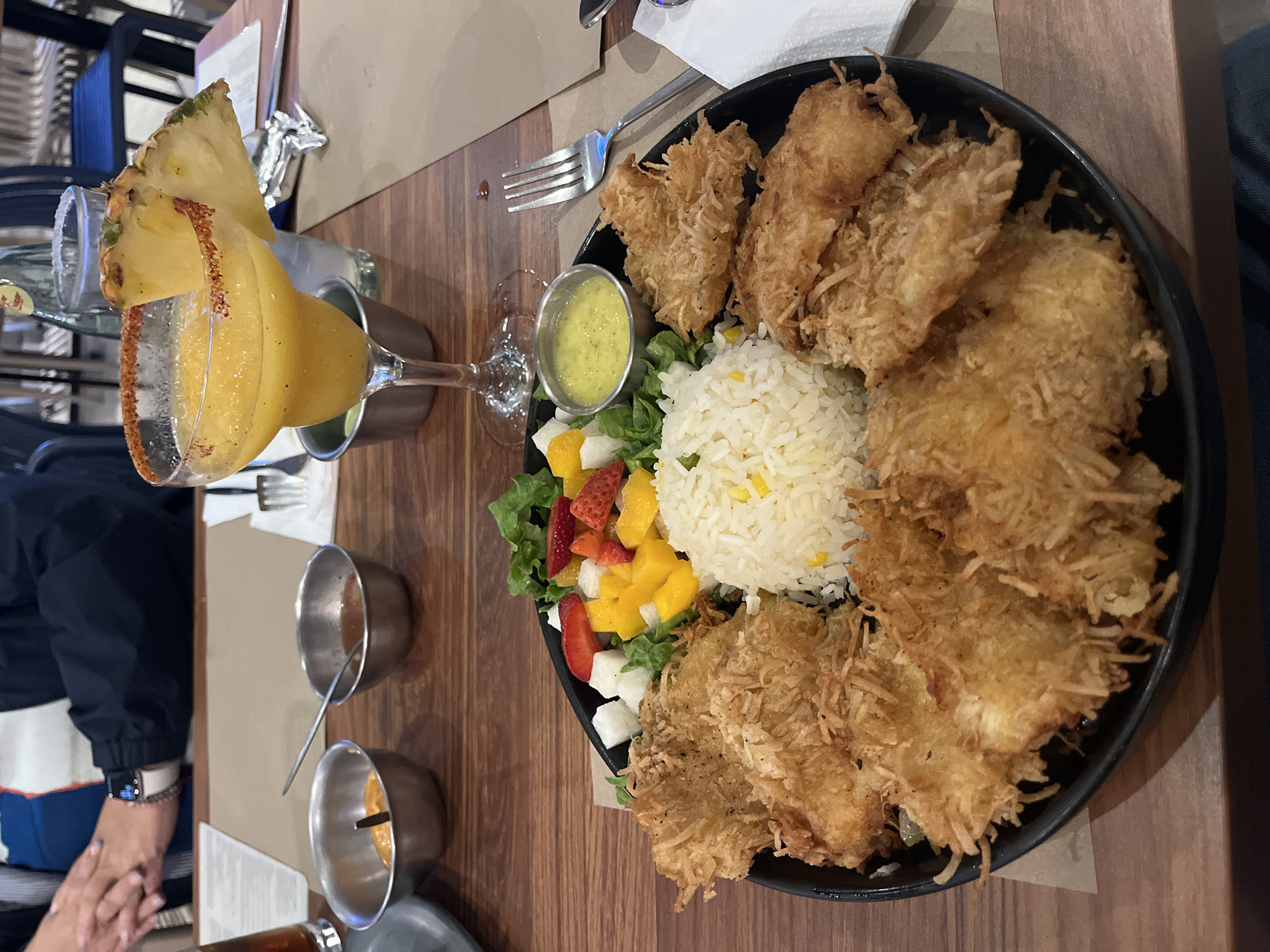 The image shows a table setting with a plate of food and a drink. The plate contains a portion of rice, a salad with mango, strawberries and white cheese cubes, fried coconut-crusted fish and a side of yellow sauce. To the left of the plate, there is a glass of orange drink garnished with a slice of pineapple. There are small metal bowls and silverware on the table as well. In the background, there is a person sitting at the table with their hands visible. The picture was taken in Puebla, Mexico.
