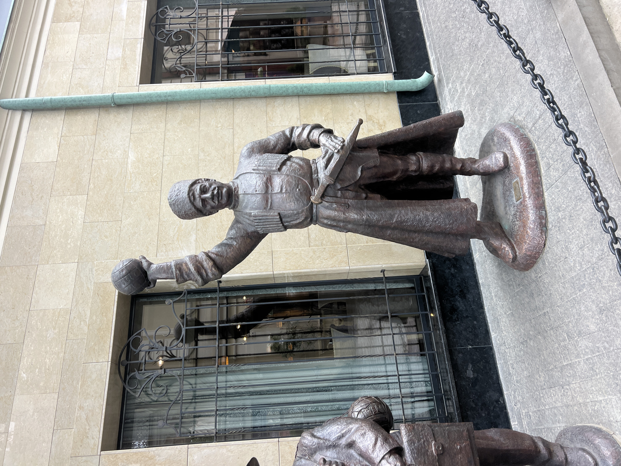 The image shows a bronze statue of a man in traditional Georgian attire, standing on a cobblestone sidewalk in front of a building with cream-colored stone walls and barred windows. The man is wearing a high-brimmed hat, a long coat, and boots, and he holds a ball in his raised right hand and a dagger in his left. Another bronze statue of a man in a suit, carrying a briefcase, is partially visible to the left. A green downspout runs along the building's facade, and a metal chain is strung across the foreground. The statue is located in Tbilisi, Georgia, and is likely part of a historical or cultural display.
