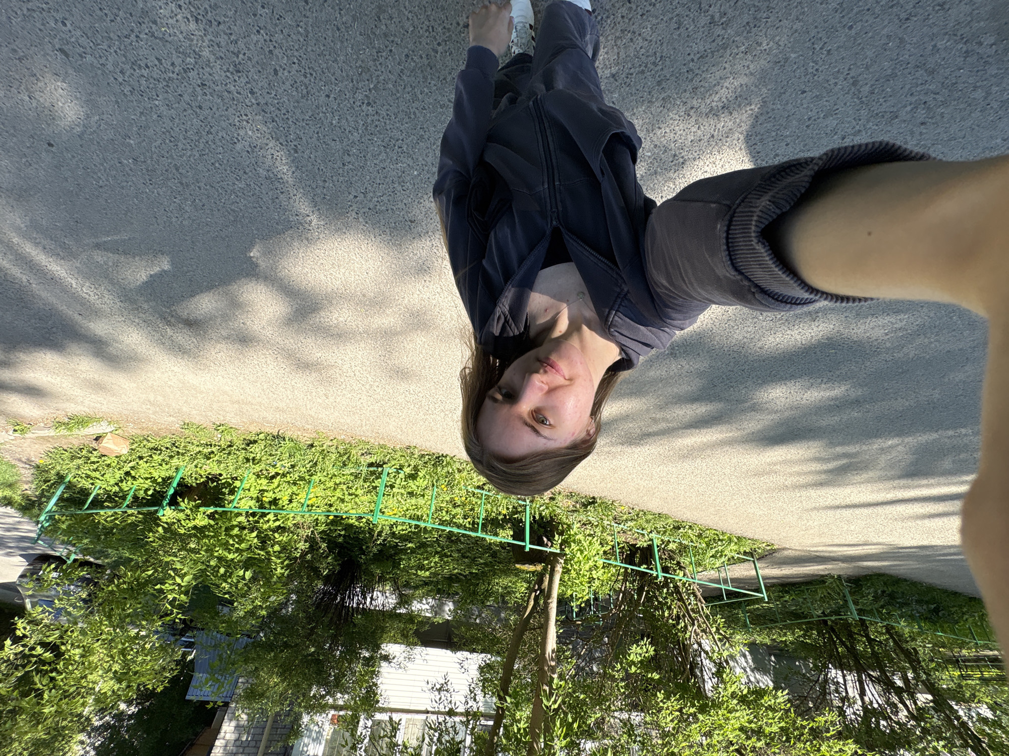 A selfie photograph shows a person with long brown hair, wearing a dark zip-up jacket and dark pants, standing on an asphalt surface, likely a street or sidewalk. The camera is held at arm's length, angled downwards. Behind the person is a green metal fence and dense vegetation, including trees and bushes. A building, possibly a house, is partially visible through the foliage. Based on the vegetation and architectural style, the location may be Perm, Russia.