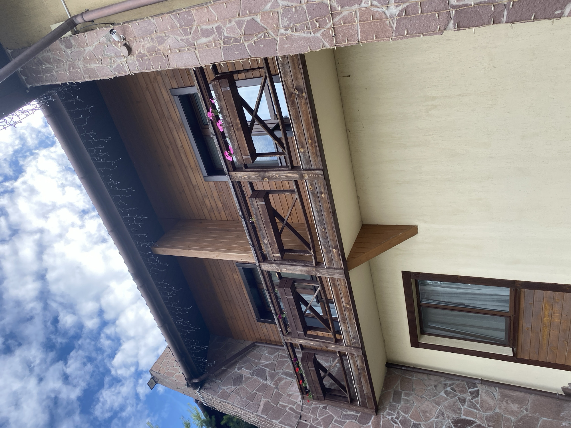 The image depicts an exterior view of a building's facade, with a focus on a wooden balcony and surrounding architectural elements. The perspective is from below, looking upwards, and the image is oriented vertically.
The building features a prominent wooden balcony with a railing constructed from thick, dark wooden beams arranged in a lattice pattern. Small planters with pink and red flowers are attached to the balcony's front, adding a touch of color. Above the balcony, the building's walls are clad in light-colored siding, contrasting with a section of reddish-brown stone masonry above the balcony.
A wooden overhang, possibly part of a roof or upper floor, extends over the scene. It is decorated with strings of small, white decorative lights, suggesting a festive or evening ambiance, though the bright daylight suggests they are not currently illuminated. A security camera is visible mounted on the underside of the overhang.
Several windows are visible. One is a dark-framed window on the wooden-clad section of the building, partially obscured by the balcony. Another is a wider, horizontally oriented window with a dark wooden frame on the light-colored wall, featuring blinds or slats.
The sky is visible in the upper left portion of the image, filled with scattered white clouds against a bright blue backdrop, indicating daytime with fair weather. The overall setting appears to be a residential or hospitality building, possibly a guesthouse or a home with rustic architectural features.
There is no visible text in the image. The time of day appears to be daytime, and the weather is clear and cloudy. The architectural style suggests a location with a focus on natural materials and a potentially cozy or traditional aesthetic.