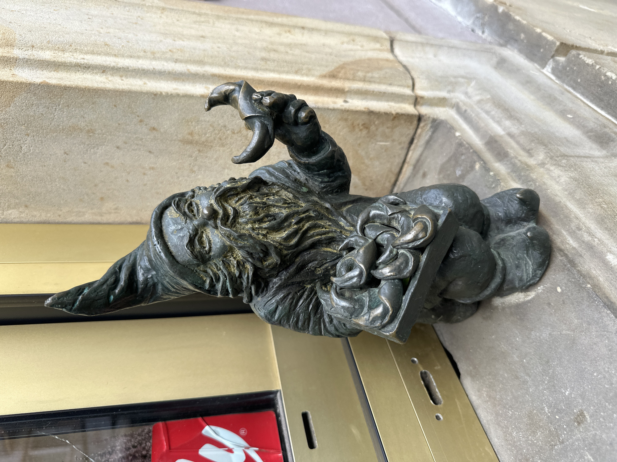 A bronze statue of a Wrocław dwarf is attached to a golden door frame near a sandstone building in Wrocław, Poland. The dwarf, wearing a cap, has a long beard, and its right hand is raised with claw-like fingers. In its left hand, it holds a square box filled with flower-like figures. Part of a red and white object is visible through a window in the door behind the statue. The sandstone wall behind the dwarf is textured and appears to be made of layered blocks.