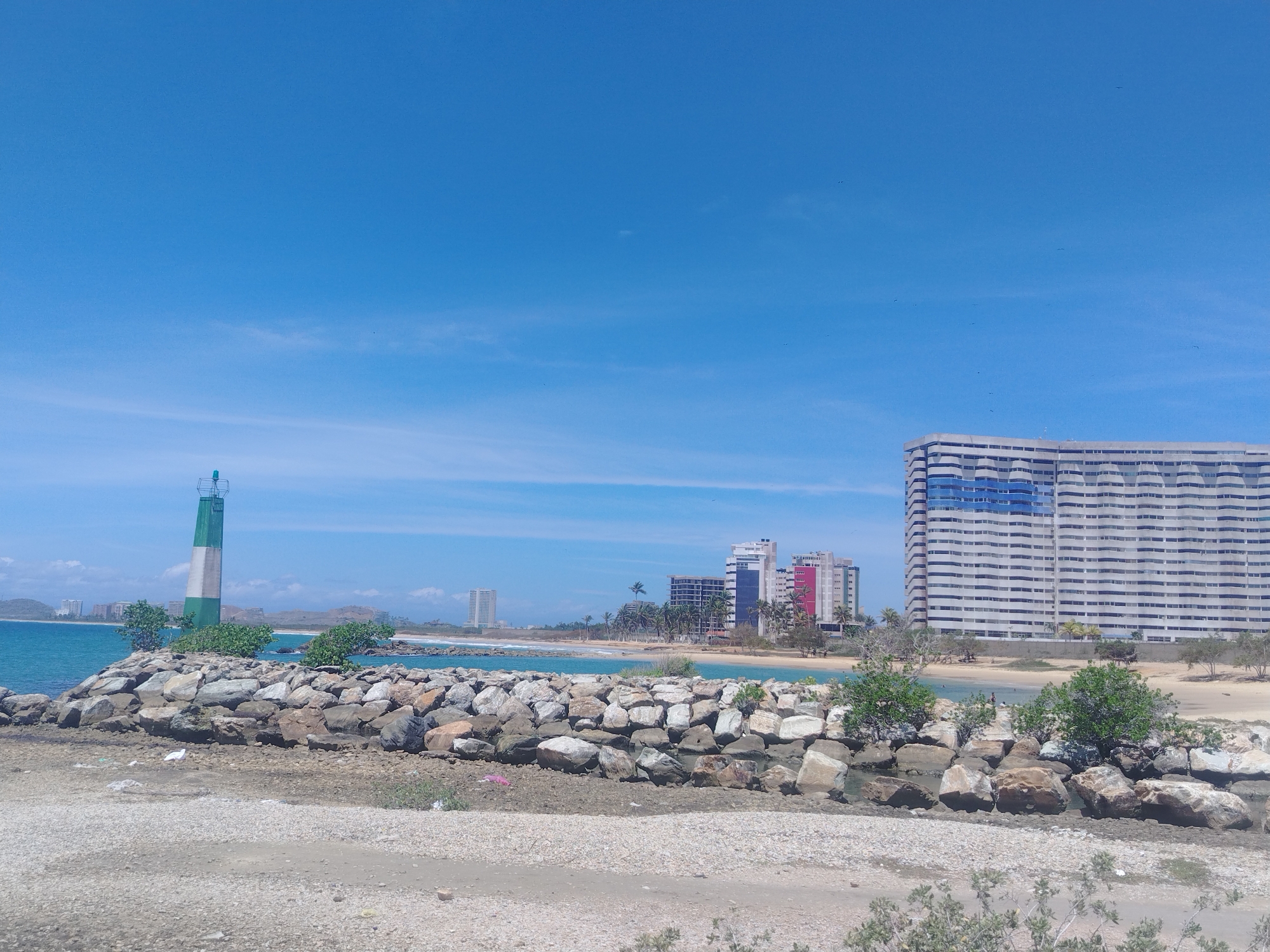 A wide-angle, eye-level shot captures a coastal scene in Porlamar, Venezuela under a clear blue sky with wispy clouds. In the foreground, a rocky breakwater juts into the turquoise ocean. Behind it, a green and white lighthouse stands prominently on the rocky shore, surrounded by small green bushes.
Further back, a sandy beach is visible, dotted with palm trees. Several modern, multi-story buildings line the coastline, including a large, white building with blue horizontal accents, and other structures with a mix of white, pink, and blue facades. The water appears calm, with a few people visible near the shore. The sun is high in the sky, suggesting it is midday. The overall atmosphere is one of a bright, sunny day at a tropical coastal resort.
