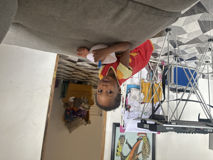 The image depicts a young child indoors in Ilishan, Nigeria. The child, with dark skin and short dark hair, is positioned in the foreground, leaning over the edge of a gray fabric sofa. They are wearing a red short-sleeved top with yellow shoulder accents and are holding a small white bowl with a blue spoon, looking directly towards the viewer.
Behind the child and to the left, a silver-colored, collapsible stand supports DJ equipment, specifically a RANE TWELVE controller and a device labeled "serato RANE." Beneath this stand, a blue hard case with silver latches rests on the floor. Further left and slightly behind the equipment, a white clothing rack is visible, laden with various light-colored garments. On the white wall behind the clothing rack, a framed abstract artwork with colorful, stylized figures is displayed.
To the right and rear, an open doorway with a wooden frame leads into an adjacent room. Through this doorway, a yellow armchair heavily covered with clothing and other items is discernible. The entire visible floor area is covered with black and white geometric patterned tiles. The walls are uniformly white, and the scene is illuminated by ambient light.