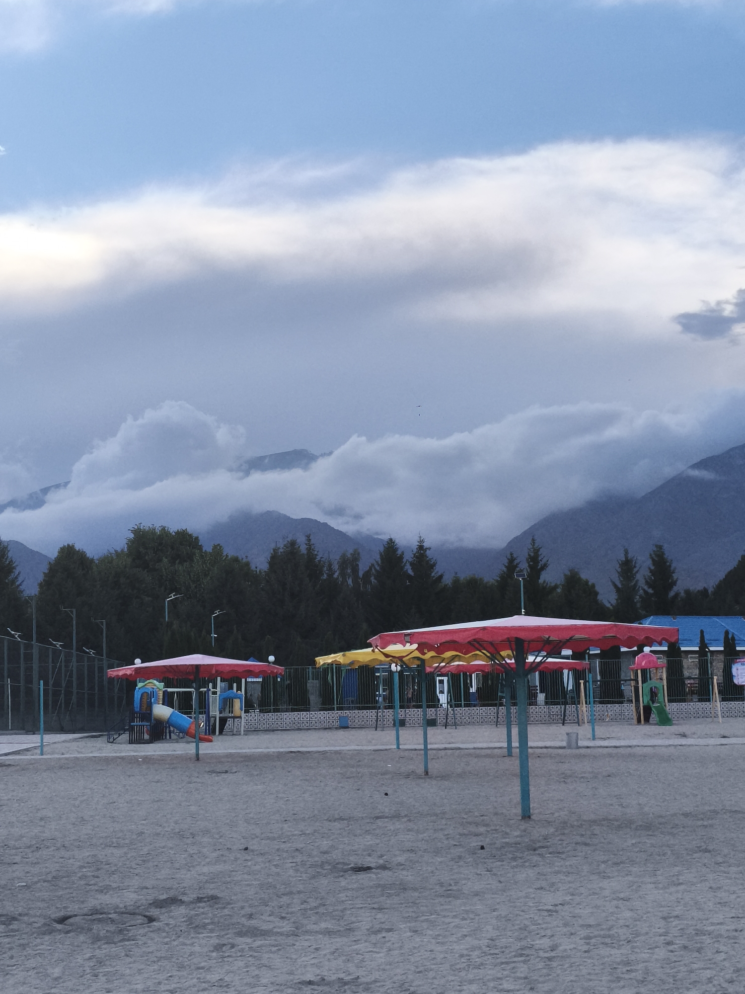 The image depicts an outdoor scene in the city of Bayet, Kyrgyzstan. In the foreground, a sandy area features several brightly colored beach umbrellas, one predominantly red and others with yellow accents. Several children's playground structures are visible behind the umbrellas, including slides and climbing frames. A dark green fence borders the playground area. Beyond the playground, a dense line of dark green coniferous trees forms a backdrop. In the distance, layered mountains rise, with their peaks partially obscured by dramatic, heavy grey clouds. The sky above is a mixture of blue and grey. The overall impression is of a recreational area situated near a natural landscape, under a somewhat overcast sky.