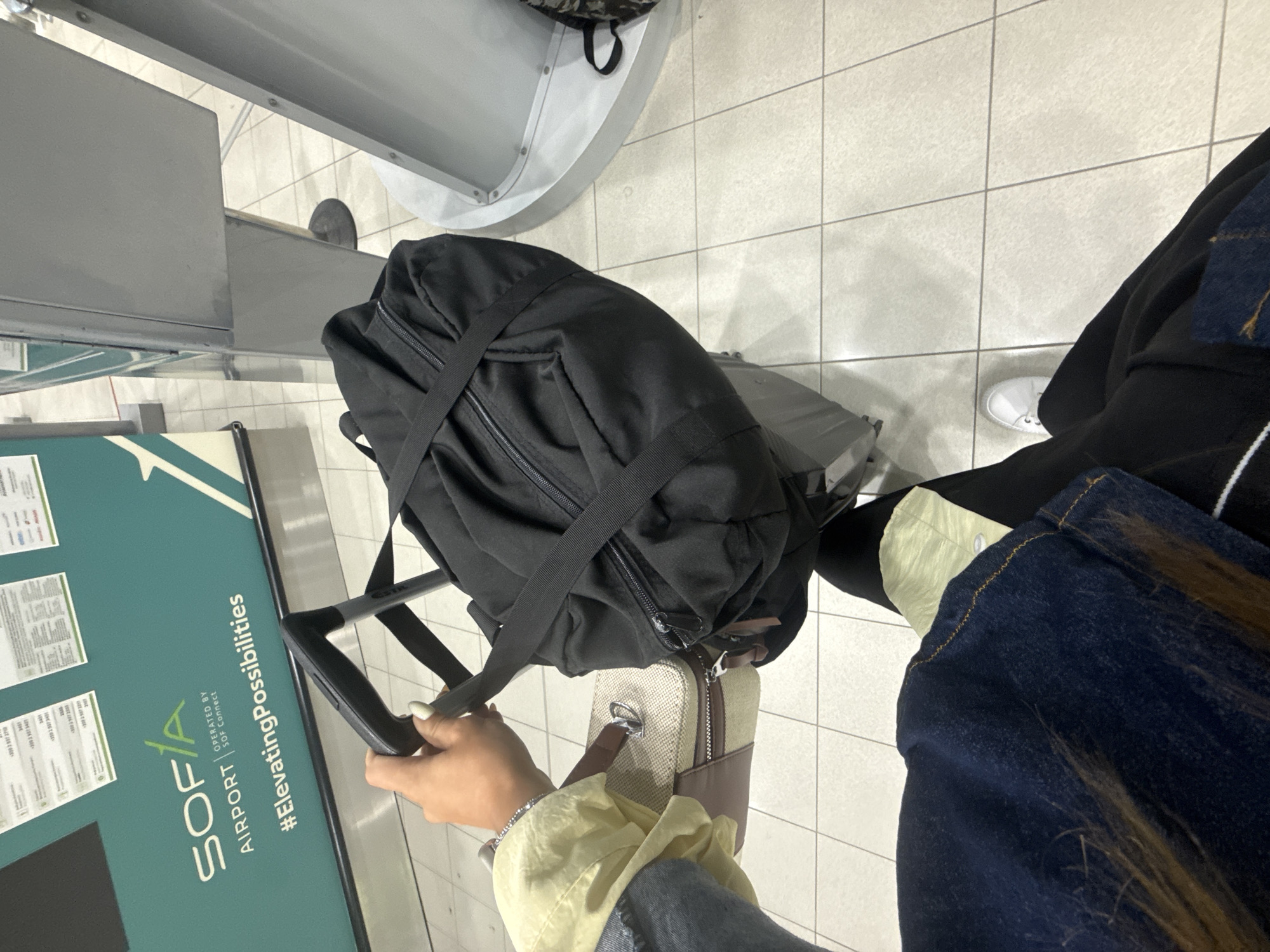 A first-person perspective shot captures a person holding the handle of a wheeled suitcase in what appears to be an airport terminal. The suitcase has a large black duffel bag strapped on top of it. To the left of the suitcase, a smaller beige and brown handbag is visible, held by the person's left hand which is also gripping the suitcase handle. In the background, a wall displays the "SOFIA AIRPORT" logo and the hashtag "#ElevatingPossibilities". The flooring is tiled.