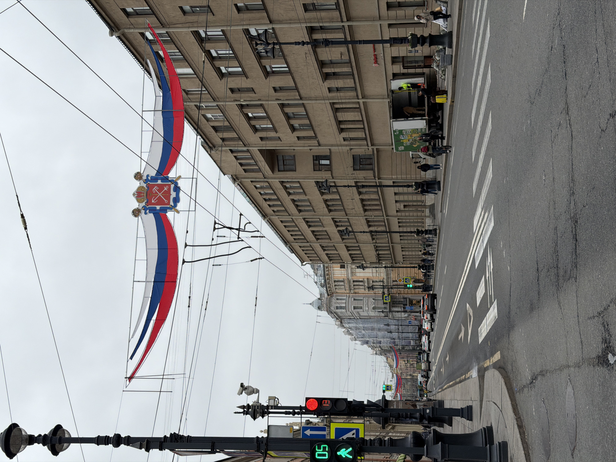 The image shows a street in Saint Petersburg, Russia. In the foreground is a black lamppost with two lamps, and a traffic light showing a red light and a green walking signal. A street sign showing "50" (presumably a speed limit) and a left-pointing arrow are visible below the traffic light. A pedestrian crossing is visible on the right side of the street. The road surface is asphalt. On the left side, hanging from cables stretched between buildings, are decorative flags with the colors white, blue, and red. A coat of arms is situated between the flags. Buildings line both sides of the street, with multiple windows and a consistent architectural style. People are walking on the sidewalk on the right side, and a truck is parked near the curb. The sky is overcast. Cars are driving down the street in the distance.