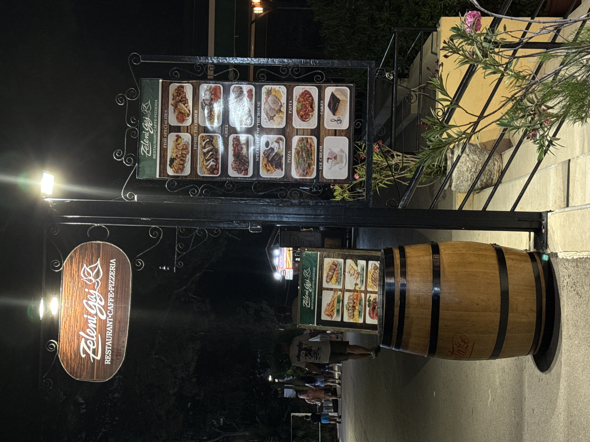 At nighttime in Budva, Montenegro, a wooden barrel serves as a planter to the left of a menu board. The menu board, which is illuminated by a light fixture above, displays pictures of various dishes and categorizes them into sections such as "Fish Specialities," "Dishes to Order," "Grill," "Specialities of the House," "Pizza," "Pasta," "Ice Cream," and "Cakes." The sign also reads "Zeleni Gaj" and "Restaurant-Caffe-Pizzeria." Further to the left, a pathway is visible with several people walking away from the camera. A few trees with dark foliage are behind them. In the background, distant lights from buildings are visible.