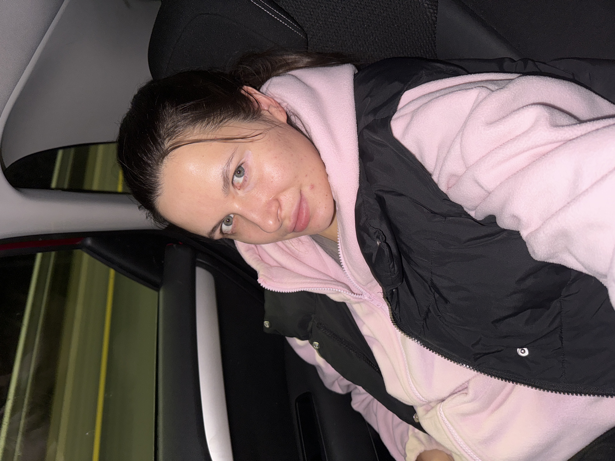 A young woman is seated in a car. She is wearing a pink hoodie under a black vest. The car window shows reflections of street lights and infrastructure. The location cannot be determined from the image.