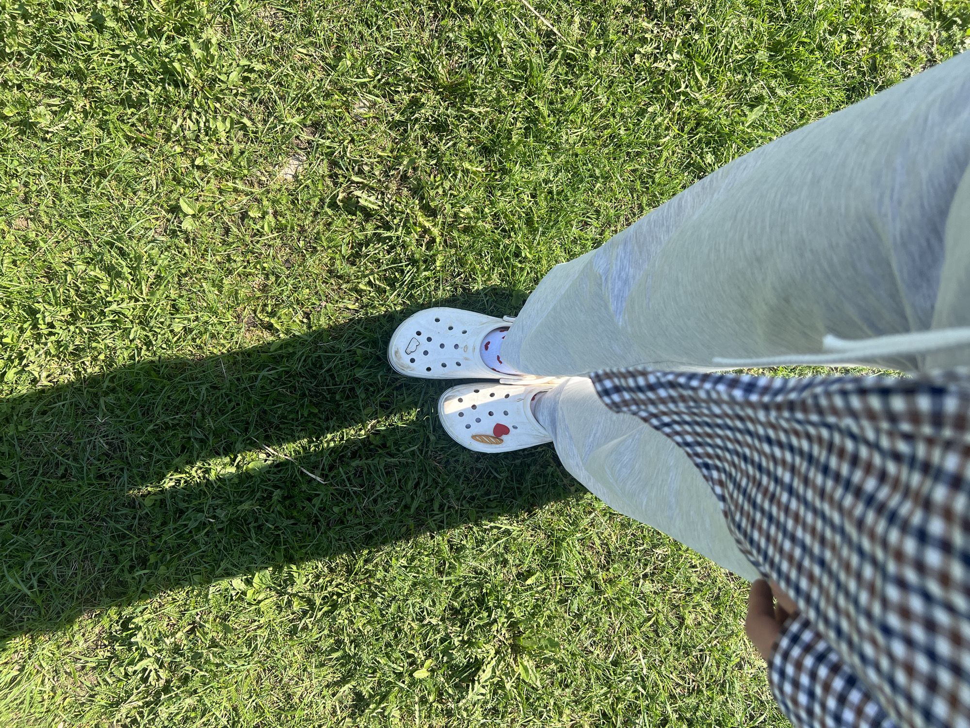 The image shows a person standing on grass in Раздолье, Russia. The person is wearing gray sweatpants, white Crocs decorated with food-themed Jibbitz charms, and socks with a red heart pattern. A brown and white plaid shirt is visible to the right. The location appears to be an outdoor grassy area.