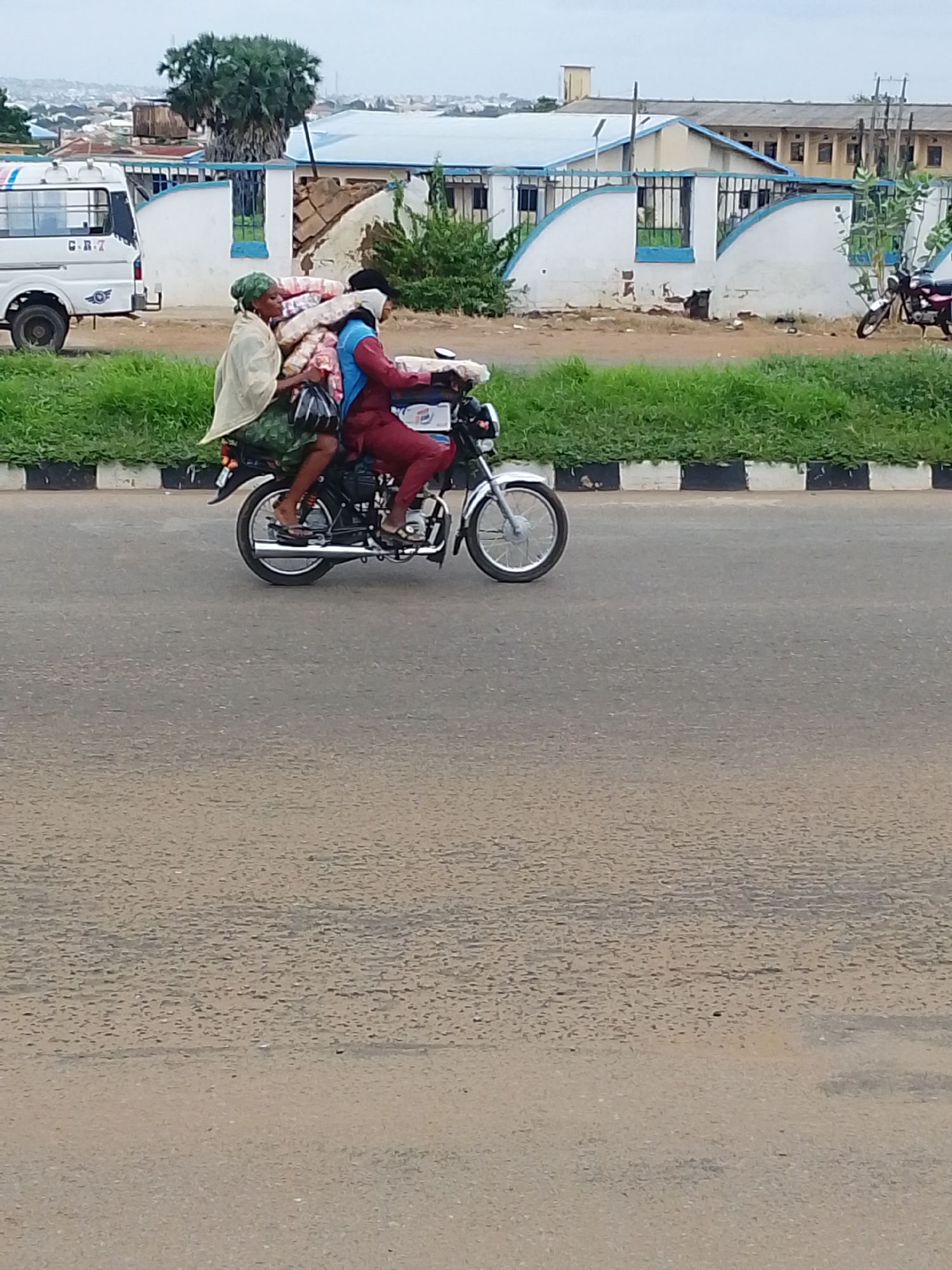 In Oja Oba, Oko Erin, Nigeria, two individuals are riding a motorcycle on a paved road. The person in front, wearing a light-colored headscarf and a patterned green skirt, is seated behind the driver. They are holding onto a large bundle of light-colored materials wrapped in a blanket. The driver, wearing a dark head covering and a red top and pants, is operating the motorcycle. The motorcycle is a dark color with silver accents, including the exhaust pipe and wheels. In the background, a white minivan is parked to the left. Beyond the road, a grassy area separates it from a white and blue building with a barred fence. Further in the distance, a cityscape with buildings and trees is visible under an overcast sky.