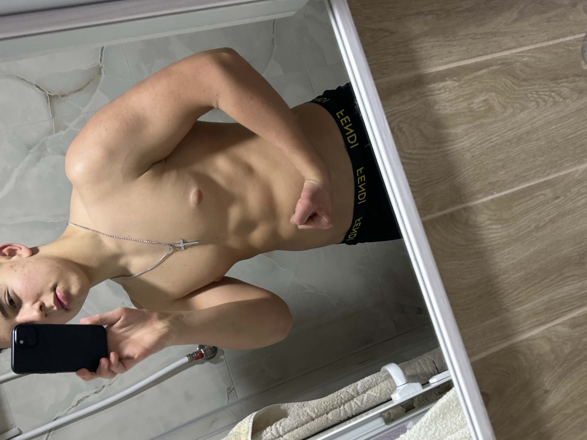A topless male is taking a mirror selfie indoors. The subject wears black underwear with a yellow Fendi logo waistband and a silver necklace with a cross pendant. He is holding a black smartphone in his left hand. The bathroom features light gray wall tiles and light wood-patterned floor tiles. A towel is visible near the bottom of the mirror frame. The location is not discernible as Zlatograd, Bulgaria.
