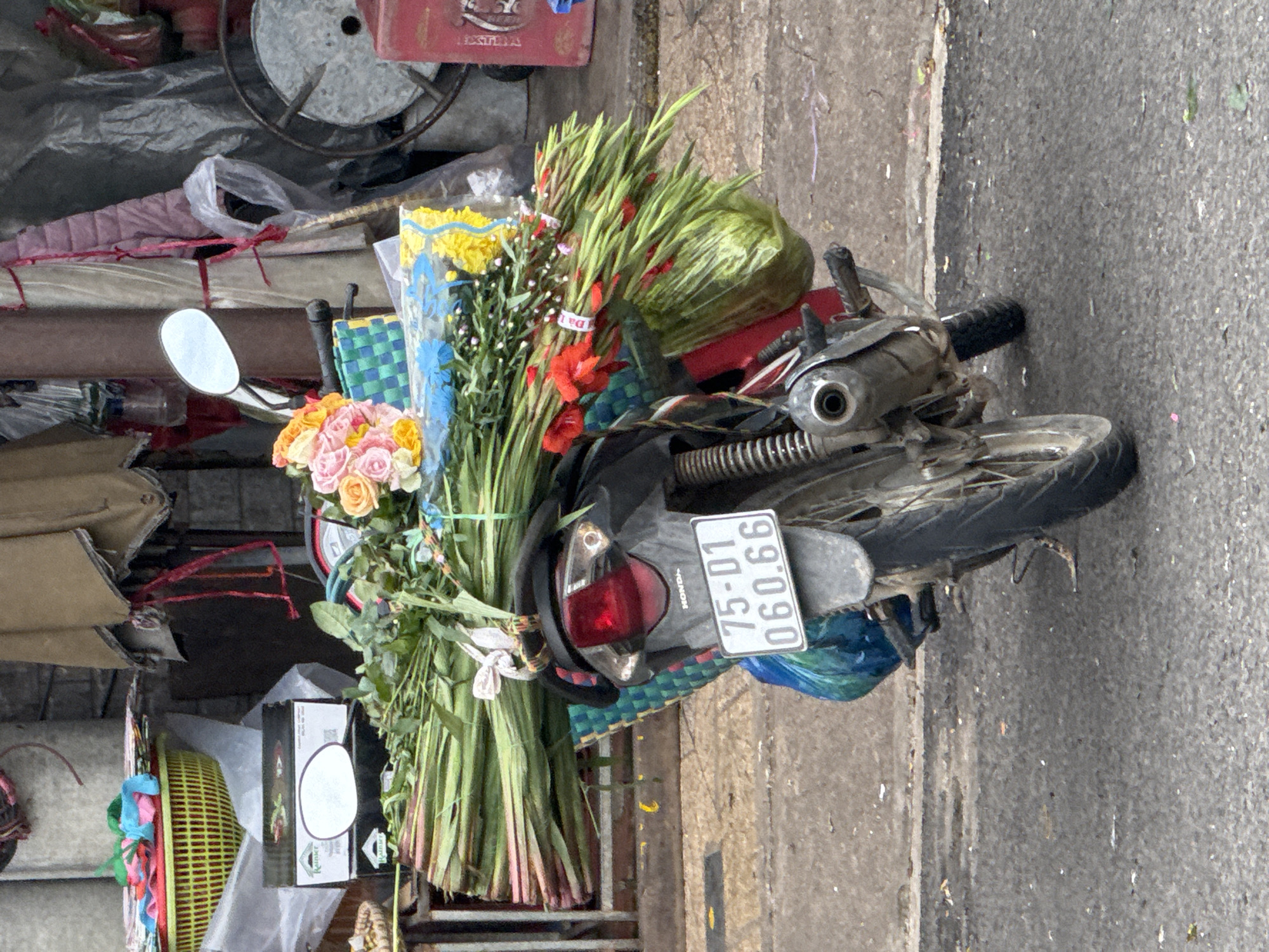 A motorcycle is parked on a paved street in Da Lat, Vietnam, loaded with a large bouquet of flowers. The motorcycle is facing away from the camera, with its rear end and license plate prominently visible. The license plate reads "75-D1 060.66". A vibrant bouquet of pink, yellow, and orange roses is tied to the back of the motorcycle, along with an assortment of green stems and red flowers. The flowers appear to be bound together and secured with twine. The motorcycle is parked in what appears to be a marketplace or street vendor area, with stacks of goods and a colorful woven basket visible in the background. The environment suggests an urban setting with a mix of commercial activity and everyday street life. The lighting indicates it is daytime.