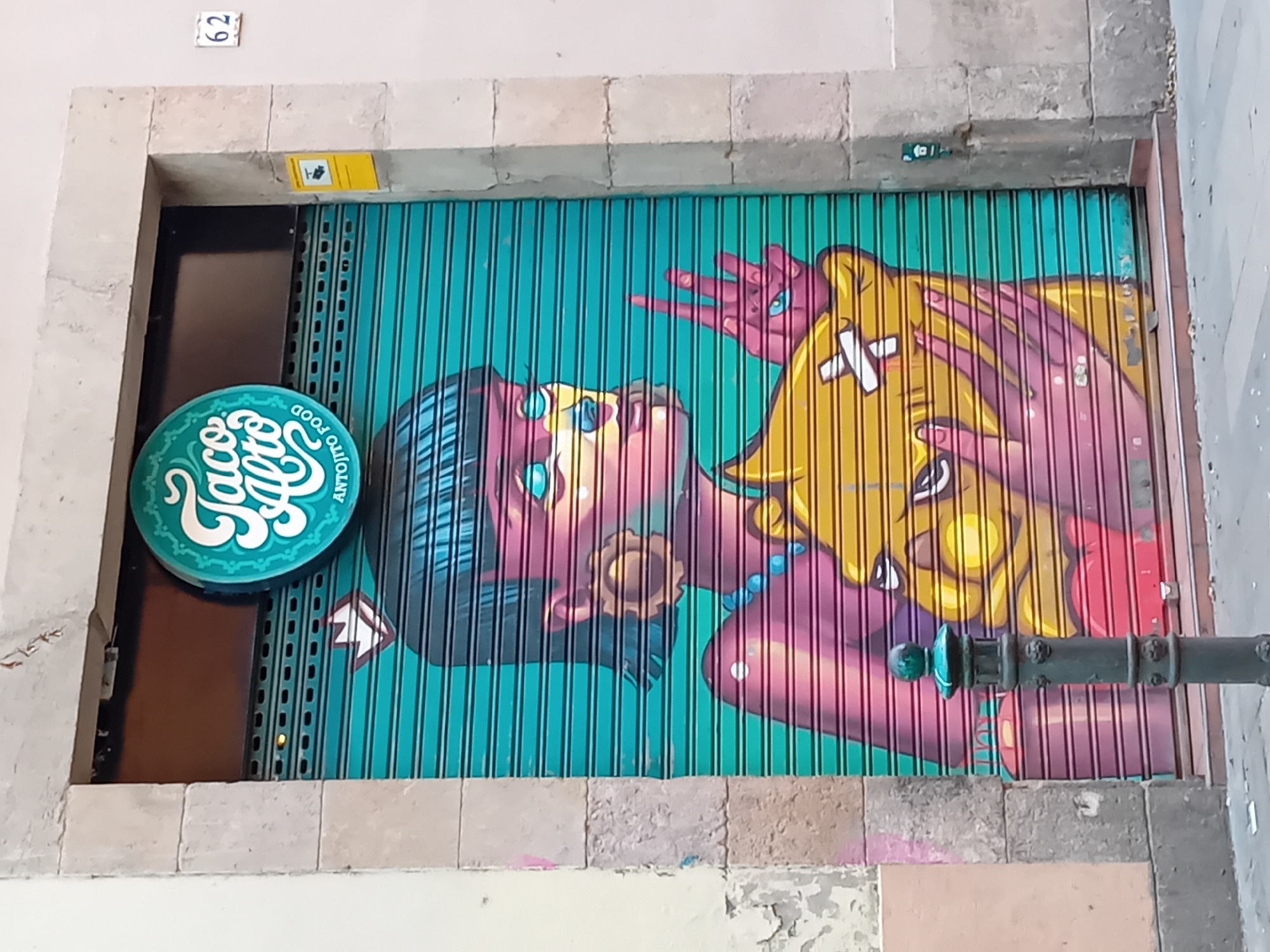 The image shows a closed metal roll-up door adorned with a mural. The mural features a stylized illustration of a woman with dark hair and a colorful face, wearing blue jewelry. She appears to be holding or interacting with a yellow object that has white "X" shapes on it. The background of the mural is a turquoise color with vertical lines.
To the left of the roll-up door is a round sign that says "Taco Alto ANTANTO FOOD" in white lettering on a green background.
The roll-up door is set into a stone or concrete wall with visible wear and tear. Above the door is a small white plaque with the number "62" on it. A metal railing is positioned in front of the mural on the right side of the image.
This image is likely taken in the city of Barcelona, Spain, based on the style of the mural and architecture in the background.