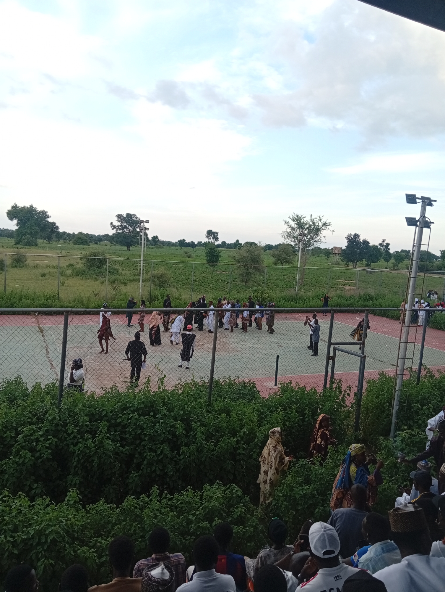 A group of people are gathered on a tennis court in Yali, Nigeria. Some of the individuals are dressed in traditional attire, while others are in more casual clothing. There appears to be an event taking place, with some people dancing and others observing. The court is surrounded by a fence, and beyond that, there is a lush green landscape with trees and fields. A tall light pole with multiple floodlights is visible on the right side of the court. The sky is partly cloudy, suggesting it may be late afternoon or early evening. The overall atmosphere seems festive and social.