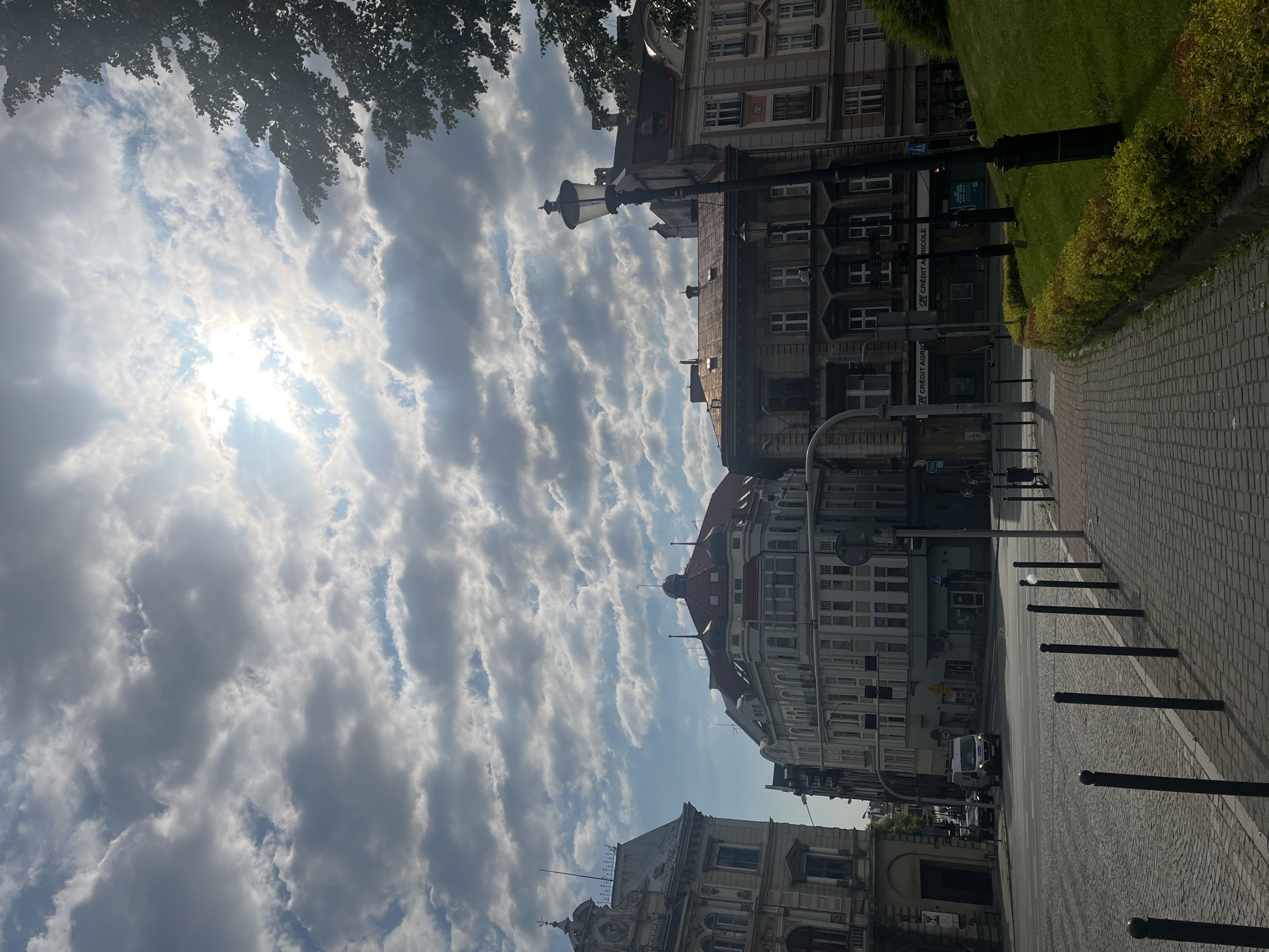 The image depicts a street scene in a European city, possibly in Poland based on the architecture and signage. The sky is partly cloudy with the sun shining through, creating bright patches and shadows. Several buildings with traditional European architecture line the street, featuring ornate facades and varied rooflines. Some buildings appear to be commercial, with visible signs for businesses. A streetlamp stands on the sidewalk, and bollards delineate the pedestrian walkway from the street. The street is paved with cobblestones, and a white van is visible driving in the distance. A grassy embankment with manicured shrubs borders the sidewalk on the right side of the image. There are no identifiable people in the foreground, but a few figures can be seen walking in the background. The overall atmosphere is that of a typical urban environment during daylight hours.