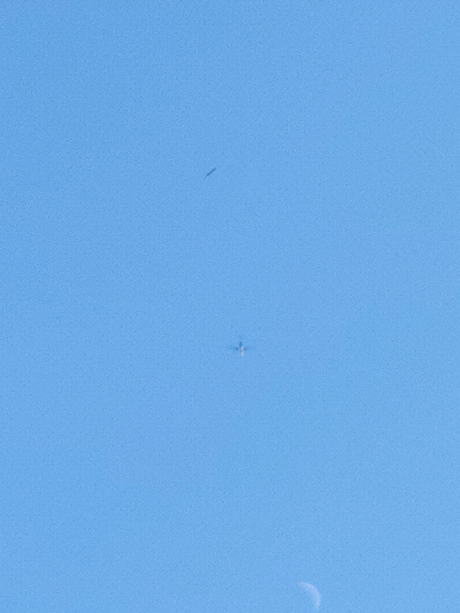 The image displays a clear, bright blue sky with a few distant objects. In the upper portion of the image, a small, dark, elongated shape is visible. Below this, a faint, cross-shaped object, possibly an aircraft, is present. In the lower right corner, a sliver of the moon can be seen. The photograph was taken in St Helens, United Kingdom.