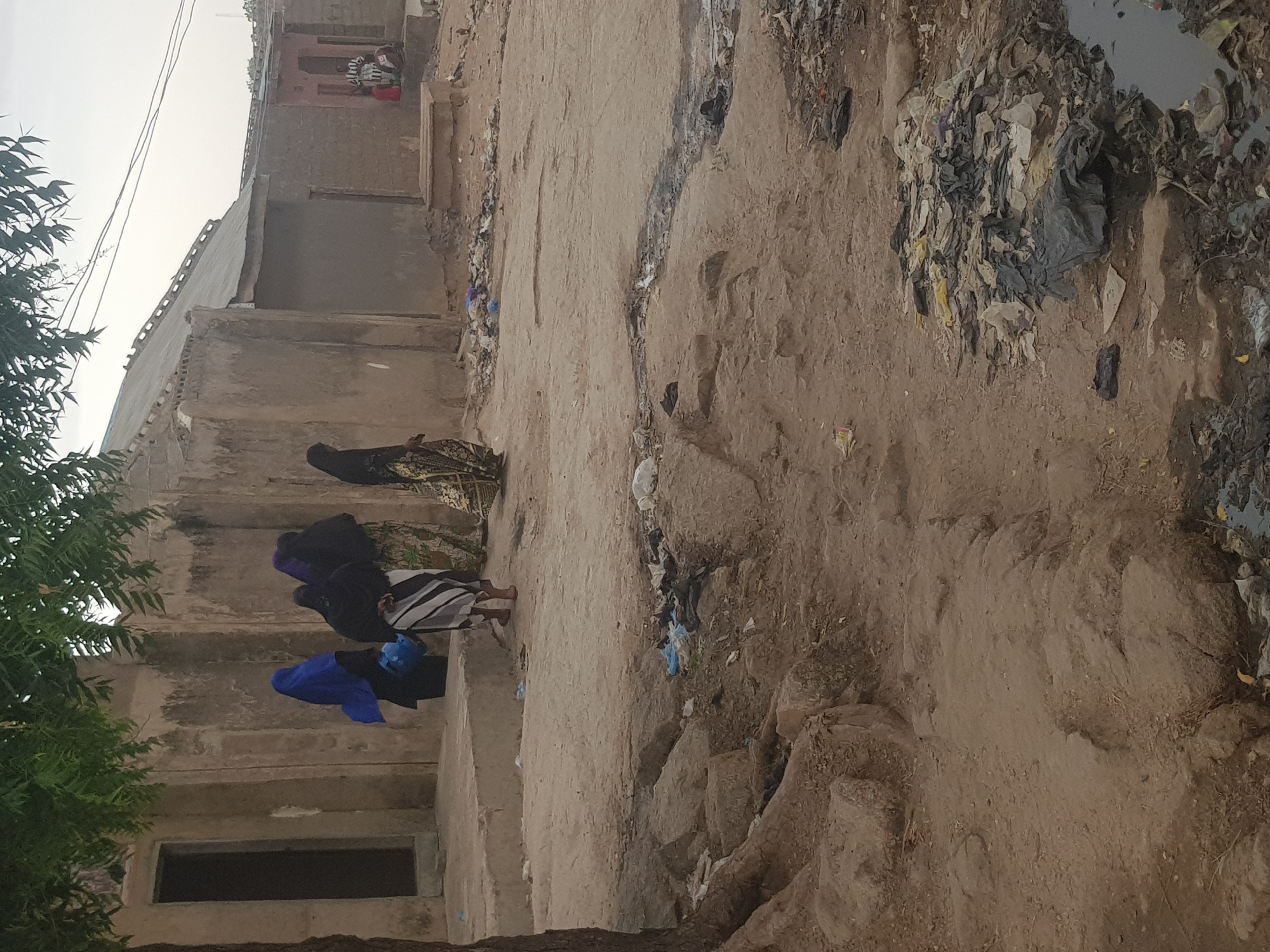 The scene depicts an impoverished area in Dabawa, Nigeria. Three women wearing traditional garments (one blue, one purple, one black) are standing near a weathered, multi-tiered concrete structure. The environment is characterized by dirt paths, debris, and accumulated trash leading towards a pool of stagnant water. Buildings in the background exhibit deteriorated facades, indicating disrepair. A person can be seen in a window.
