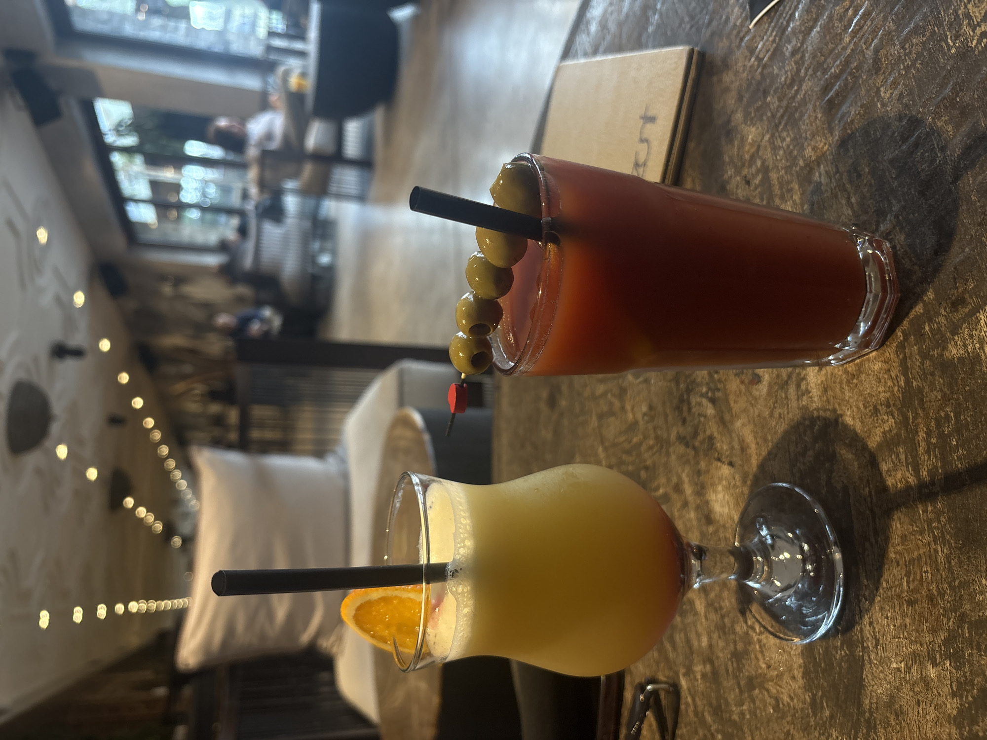 The image captures a scene within a restaurant or bar, potentially located in Varna, Bulgaria. Two drinks are prominently displayed on a textured wooden table. One is a tall, reddish-brown cocktail garnished with olives on a skewer and a black straw, likely a Bloody Mary. The other drink, in a stemmed glass, is a layered yellow and orange cocktail with a black straw and an orange slice garnish, resembling a tropical drink. Behind the drinks, the out-of-focus background shows glimpses of a well-lit interior with seating arrangements, including sofas and chairs, and strung decorative lights. Patrons are visible in the background. A menu or bill is situated behind the Bloody Mary.