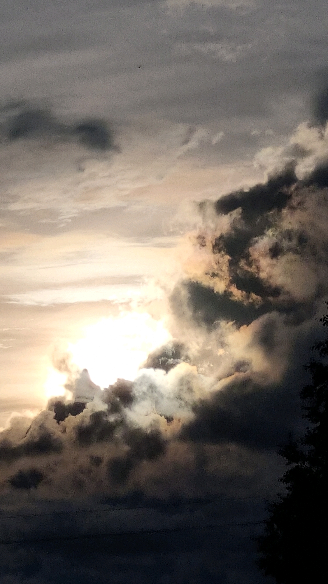 The image shows a dramatic sky with dark, heavy clouds illuminated by a bright light source, likely the sun, peeking through from the left. The light creates a striking contrast with the dark clouds, casting colorful hues of yellow and orange. The lower portion of the image is darker, possibly depicting the ground or silhouetted trees on the right. There is a small, dark object visible in the upper center of the sky, which could be a bird or an aircraft. The scene appears to be set in Vysokoe, Russia, based on the provided context.