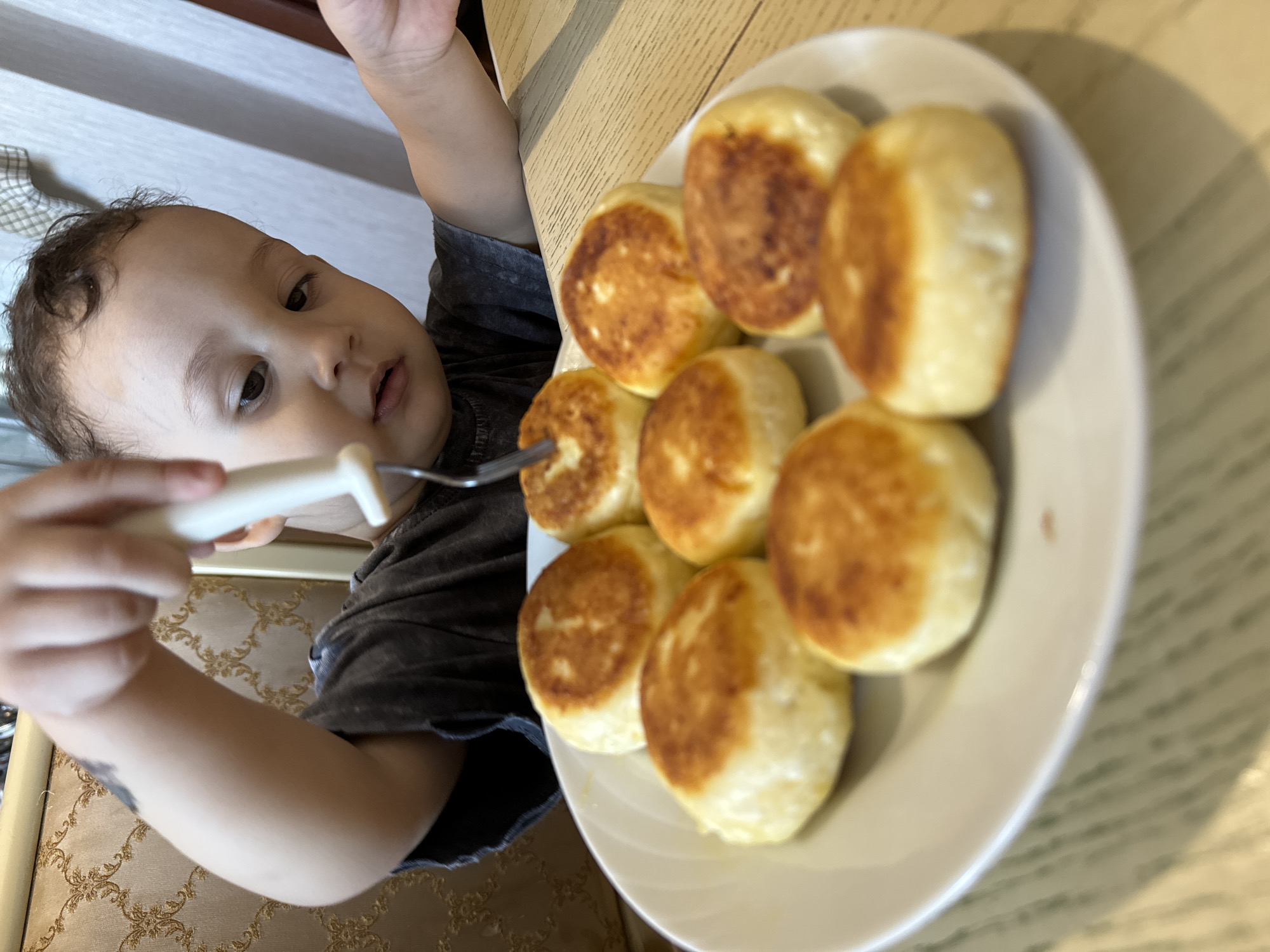 A young child holds a fork over a plate of round, browned fritters or cheese pancakes, possibly syrniki, on a white plate on a wooden table. The child has dark hair, dark eyes, and wears a dark-colored t-shirt. The scene is indoors with light from an unseen source. The location is the city of Chaikovsky, Russia.