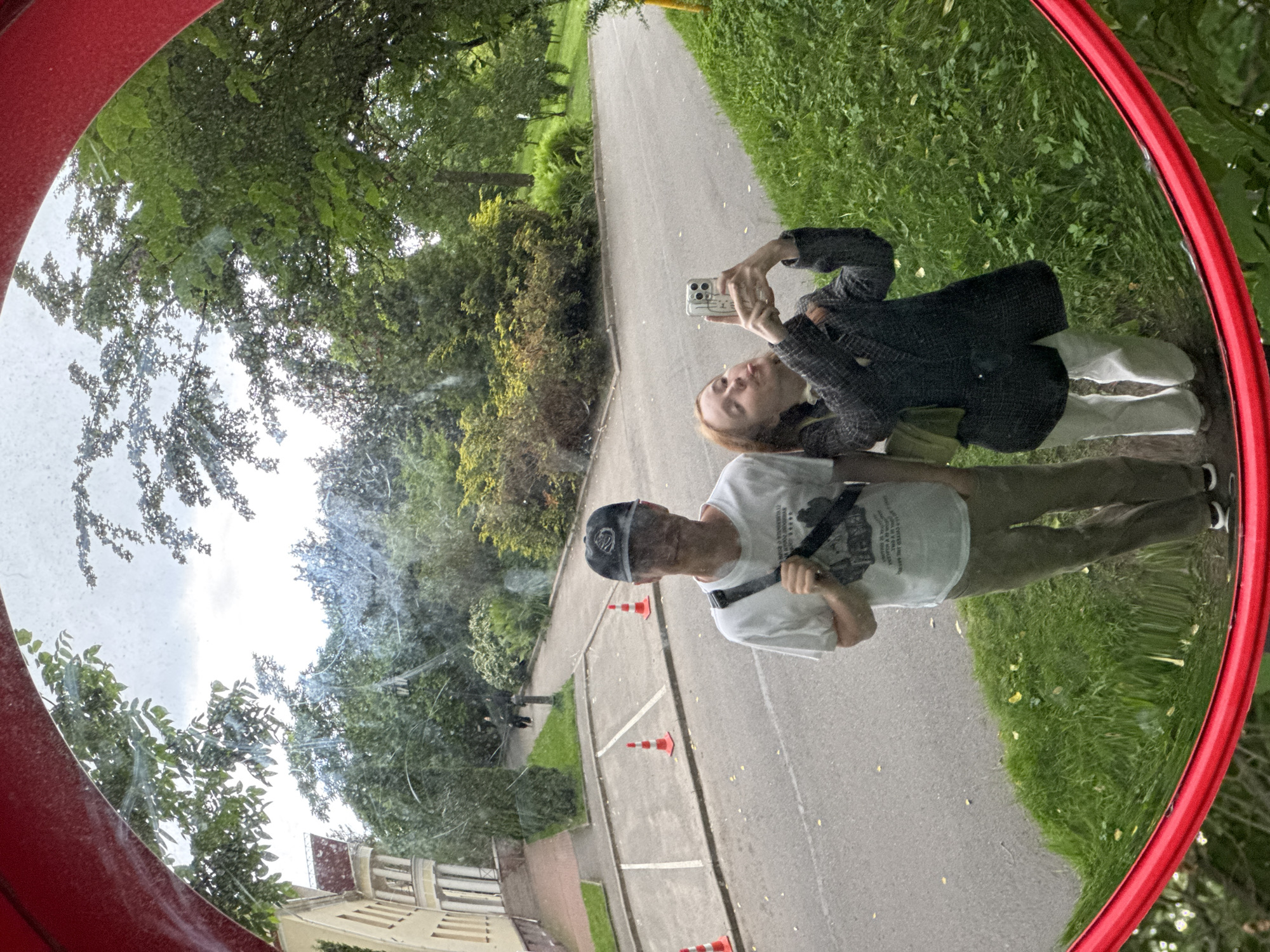 A fisheye lens captures a reflection in a convex red-rimmed mirror, showing two individuals taking a selfie in an outdoor setting that appears to be a park or university campus in Saint Petersburg, Russia. The person on the left, a male, wears a white t-shirt, khaki pants, and a black baseball cap. He has a black bag slung across his chest. The person on the right, a female, is dressed in a grey patterned jacket and white pants, holding a smartphone to take the photo. She is sticking out her tongue.
The background reflected in the mirror reveals a paved pathway, a grassy embankment, trees, and a building. The building, situated to the left, is light-colored with a red-tiled roof and classical architectural elements. Orange traffic cones are visible on the paved area. The sky above is overcast. The presence of the building and the manicured grounds suggests a location like the Smolny Institute or another campus in Saint Petersburg.