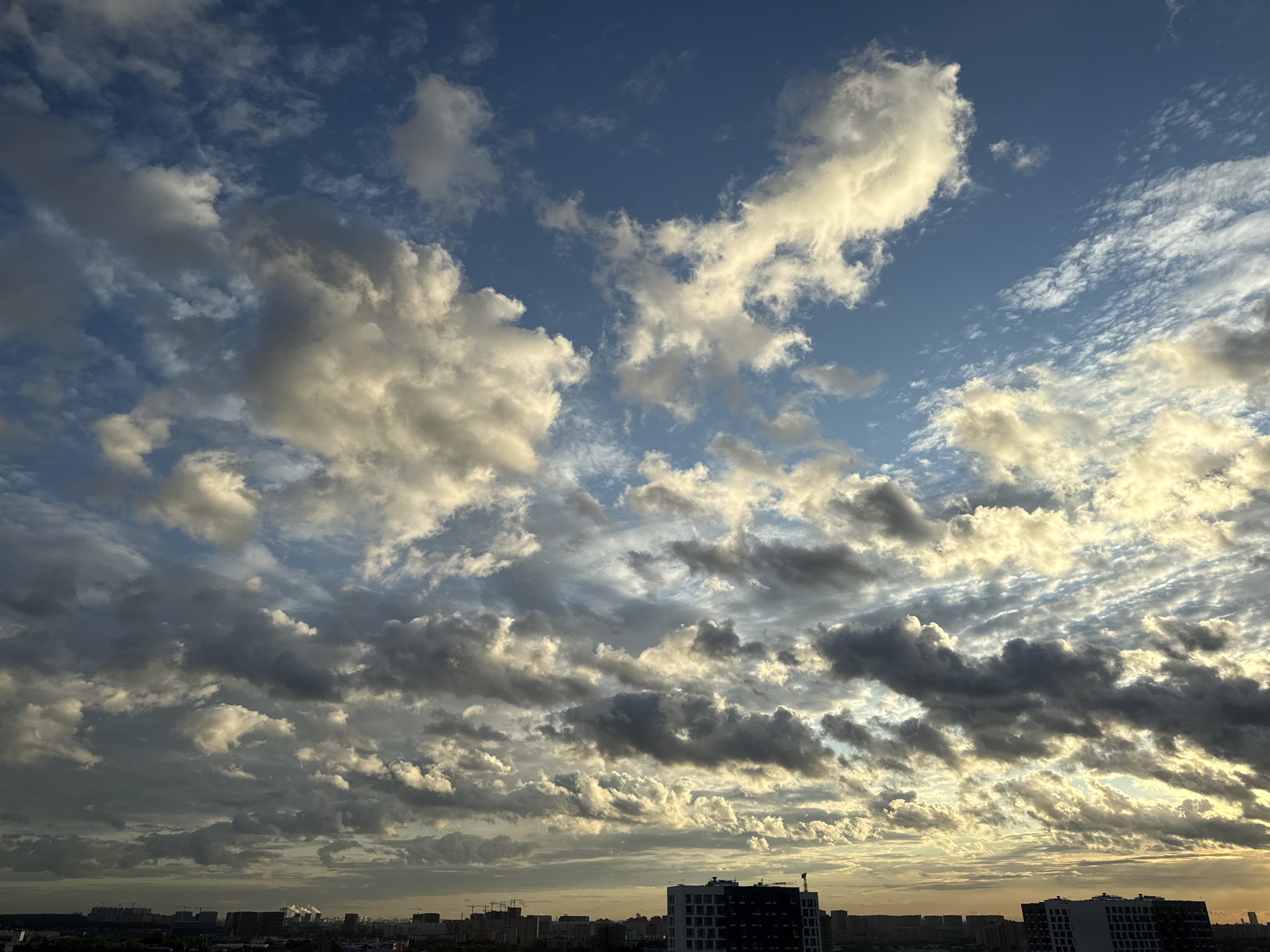 A wide-angle shot captures a dynamic sky over the city of Lyubertsy, Russia. The sky is filled with a variety of clouds, ranging from fluffy white cumulus formations to darker, more diffused stratus clouds. Sunlight pierces through gaps in the clouds, creating a dramatic play of light and shadow. The lower portion of the image reveals a cityscape with buildings of varying heights and construction stages. Some buildings are modern, with dark and white facades, while others appear to be under construction with cranes visible. The horizon line is low, emphasizing the vastness of the sky. The overall scene suggests either sunrise or sunset due to the warm, golden hues present in some of the clouds and the soft light illuminating the city. There are no people or discernible actions depicted in the image. The environment is urban, with a dense collection of buildings and what appears to be some distant industrial activity indicated by plumes of smoke.