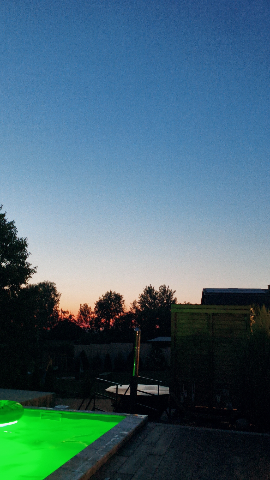 The image depicts an outdoor scene at dusk. In the foreground, a section of a swimming pool with bright green illuminated water is visible. The pool is bordered by a wooden deck and stone coping. In the middle ground, silhouetted trees line the horizon, with the sky transitioning from a clear blue to hues of orange and pink, indicating sunset. Beyond the trees, a dark wooden fence and a building with a dark roof are partially visible. Further into the background, shadowy figures of trees and structures are discernible. The overall atmosphere is tranquil and serene, characteristic of an evening in a suburban or rural setting. The image does not contain any people, specific landmarks, or identifiable features that would allow for precise location identification within Bezliudivka, Ukraine.