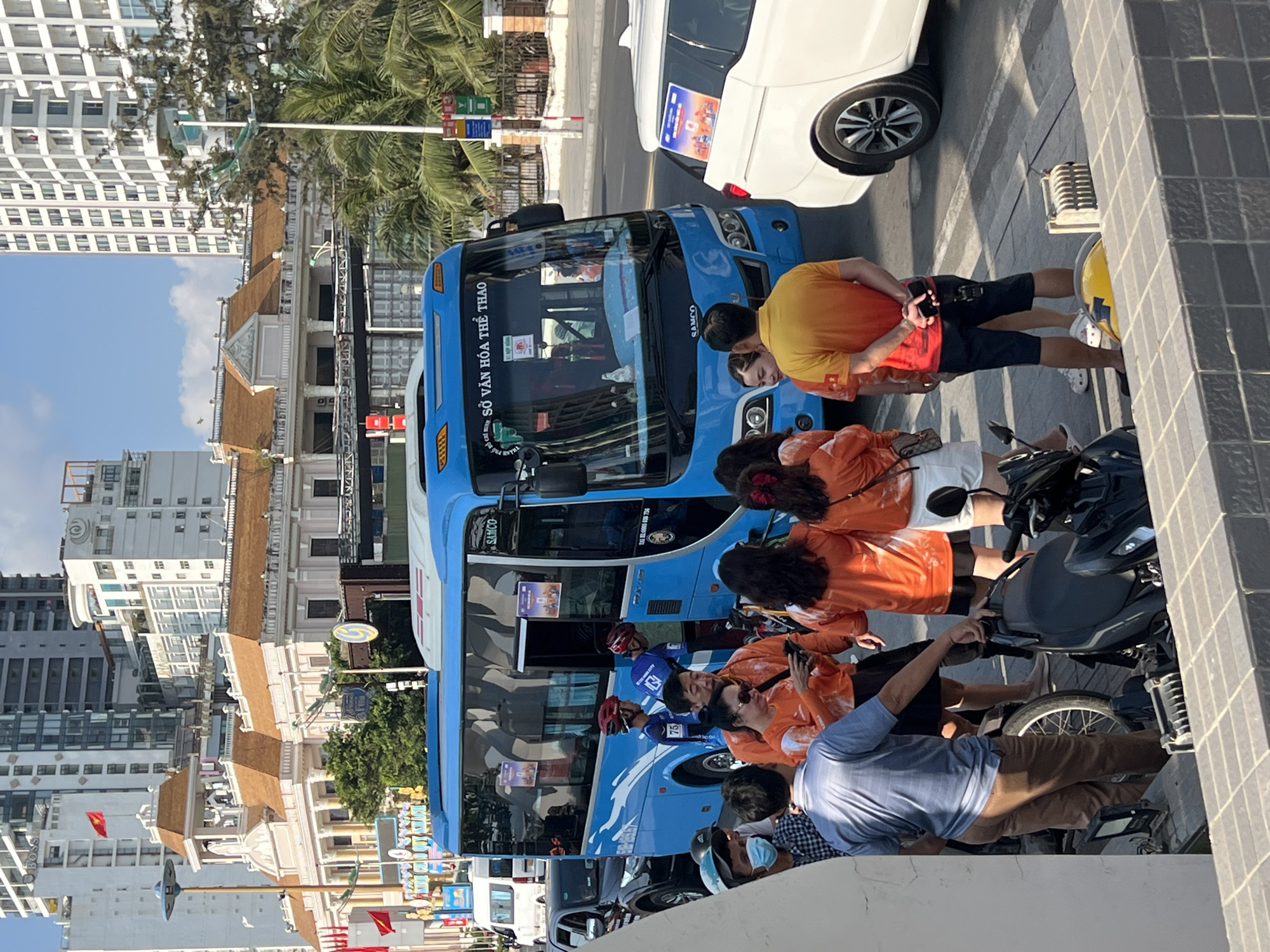 The image captures a street scene in Nha Trang, Vietnam. A blue bus labeled "SỞ VĂN HÓA THỂ THAO" (Department of Culture and Sports) is parked along the road. Several people, many wearing matching orange shirts, are standing around the bus. There are motorcycles parked near the curb as well. A white car is parked in front of the bus. The architecture in the background suggests an urban environment with high-rise buildings and traditional Vietnamese-style structures. People are seen using their mobile phones. There is a tall hotel in the background.