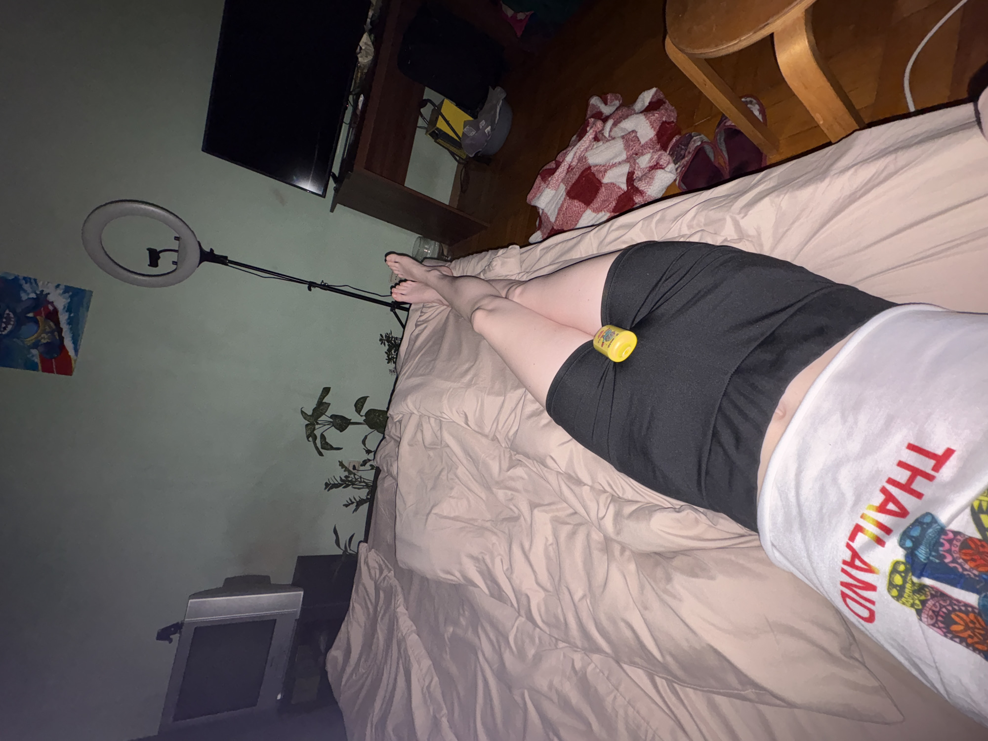 A room interior is seen from an elevated angle. A person is lying on a bed with a light pink sheet. The person is wearing black shorts and a white t-shirt with "THAILAND" printed vertically in red and blue with images of flip-flops. A yellow container sits on the person's right thigh. The person's legs are stretched out on the bed with bare feet. A ring light is on a stand near the head of the bed. In the background are a TV and entertainment center, a plaid blanket, a wooden chair, a plant, and a smaller TV. The location is unknown.