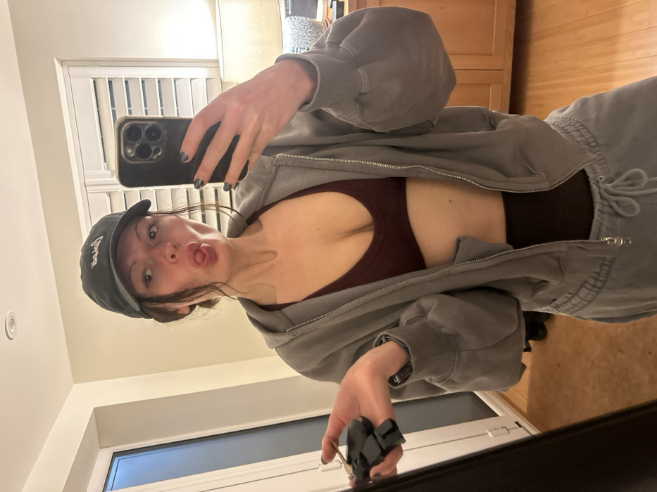 A woman poses in a mirror selfie, sticking her tongue out. She wears a dark baseball cap, a burgundy athletic top, and a gray zip-up hoodie with matching sweatpants. The background includes a window with white blinds, light-colored walls, and a wooden cabinet. She holds a phone with a black case in one hand and keys attached to a carabiner in the other. The image is slightly tilted.