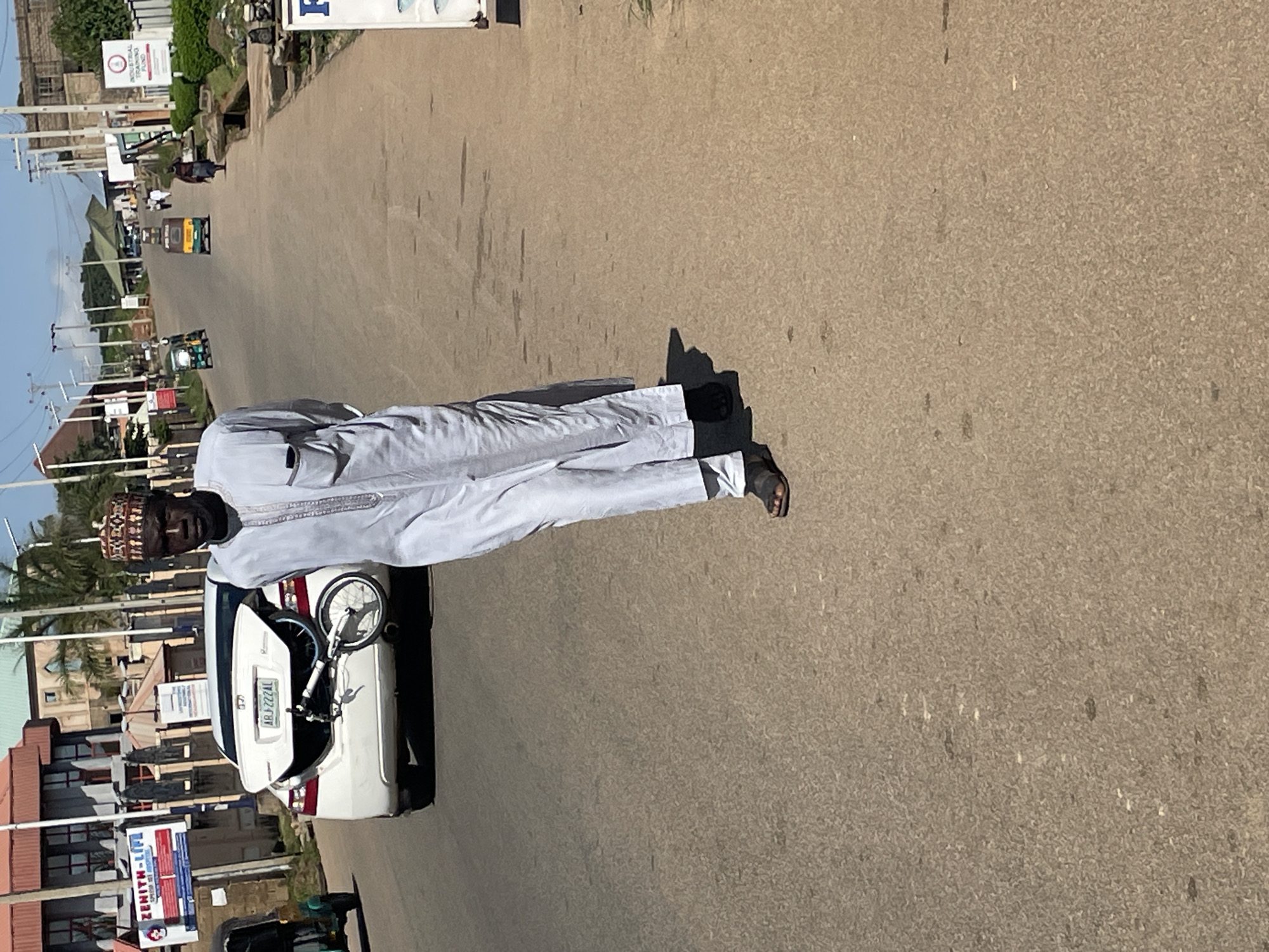 A man in traditional Islamic attire walks on a paved road in Jos, Nigeria. He wears a white flowing robe and a decorative cap. He is walking away from the camera, with his back partially turned. In the background, a white car with its trunk open is parked on the side of the road, with a disassembled bicycle inside. Other vehicles, including a tricycle-like vehicle and a few other cars, are visible further down the road. Buildings and shops line the street, and utility poles with wires are present. The sky is clear and blue.