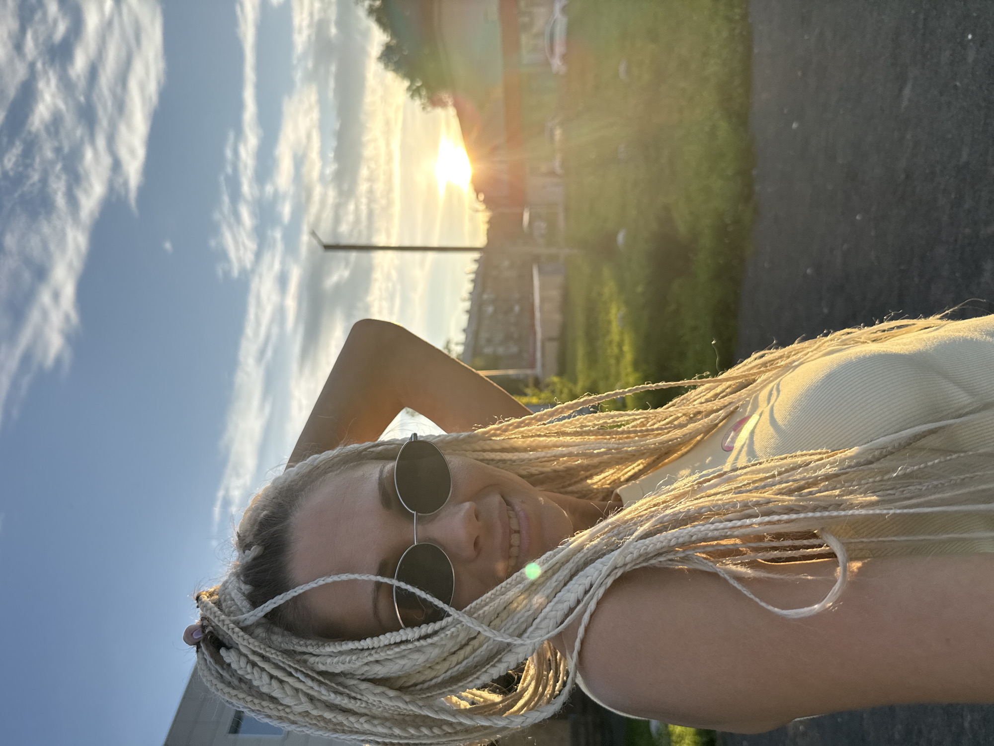 A woman with blonde box braids, wearing a light yellow tank top and oval sunglasses, smiles at the camera. She has her left hand raised behind her head. The image is a selfie taken outdoors during golden hour. The background shows a street, a grassy area, some buildings, a lamppost, and a bright sunset. The location is identified as Rusanovo, Russia.