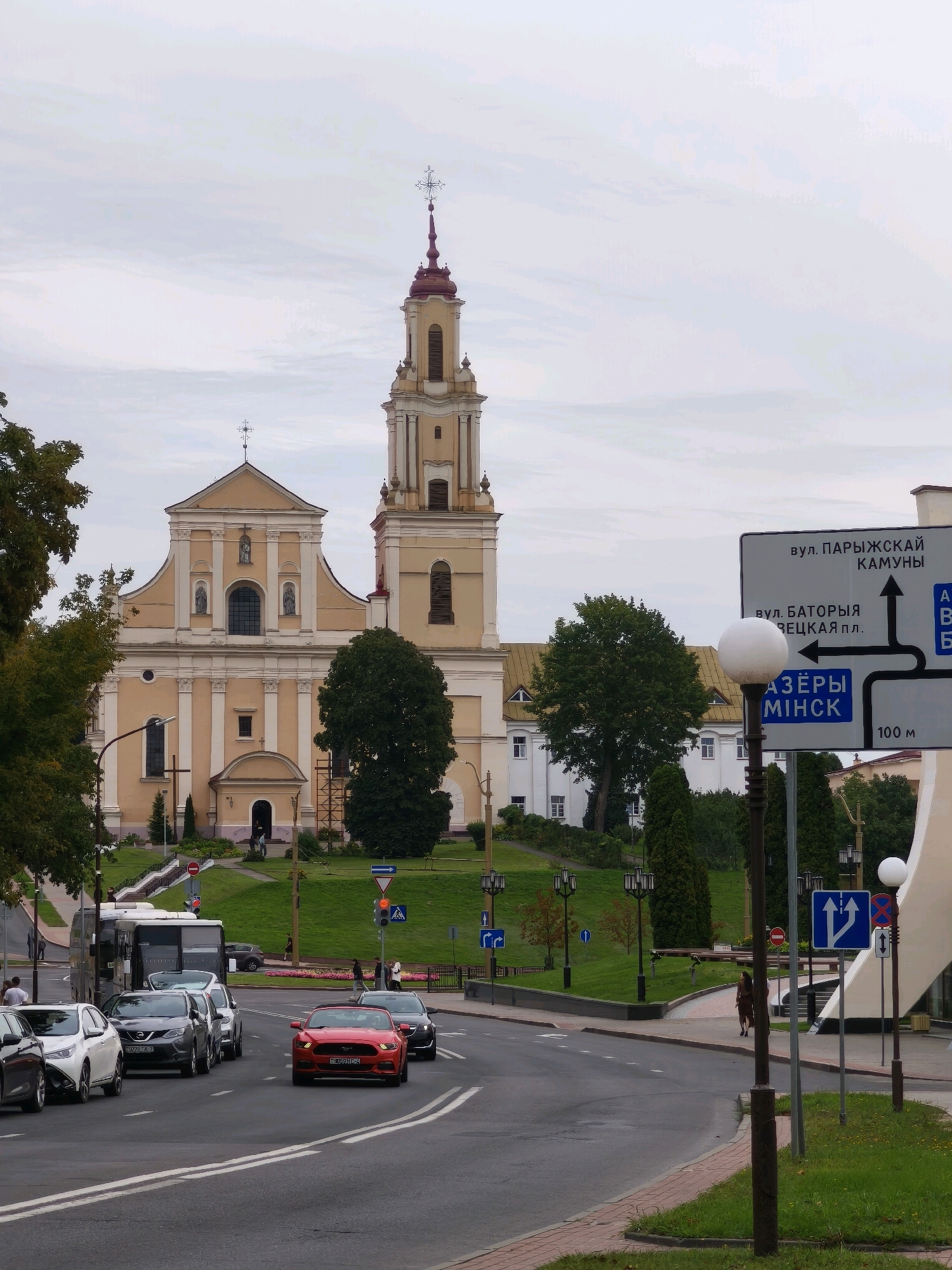 The image depicts a street scene in Hrodna, Belarus, dominated by a large, light-yellow church with a tall, prominent bell tower. The church features intricate architectural details, including arched windows, columns, and crosses atop its spires. In the foreground, a multi-lane asphalt road curves to the right. Several cars, including a red sports car, a white sedan, and a grey SUV, are driving on the road. A white bus is parked on the left side of the road. Pedestrians are visible on the sidewalk and near the church entrance. Streetlights and a traffic signpost are also present. The environment is characterized by a cloudy sky, green grass, and trees. The traffic signpost displays directions in Cyrillic script, indicating street names such as "вул. ПАРЫЖСКАЙ КАМУНЫ" and "вул. БАТОРЫЯ ЗЯЦКАЯ ПЛ.".
