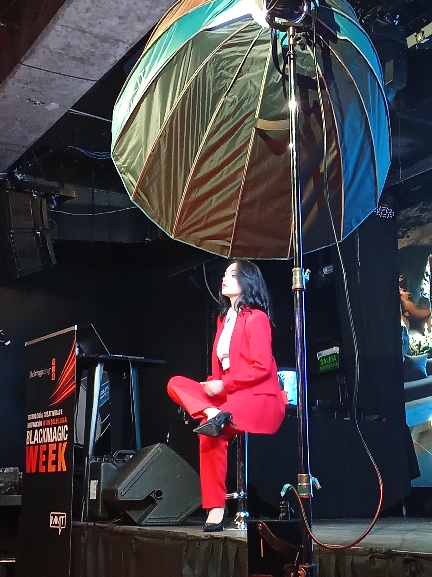 A woman in a red suit and white blouse sits on a raised platform in front of a dark background, illuminated by a large, multi-colored umbrella-like light diffuser positioned above and behind her. The diffuser has sections of green, blue, and brown. She is facing to her left, looking upwards. To her left, there is a black podium with a laptop on it, and a sign that reads "Blackmagic Week". To her right, there is a black emergency exit sign with green lettering that reads "SALIDA DE EMERGENCIA". She is wearing dark shoes. The scene appears to be indoors, likely a stage or event space. The image was taken at the Movistar Arena in Santiago, Chile, during the Blackmagic Week event.
