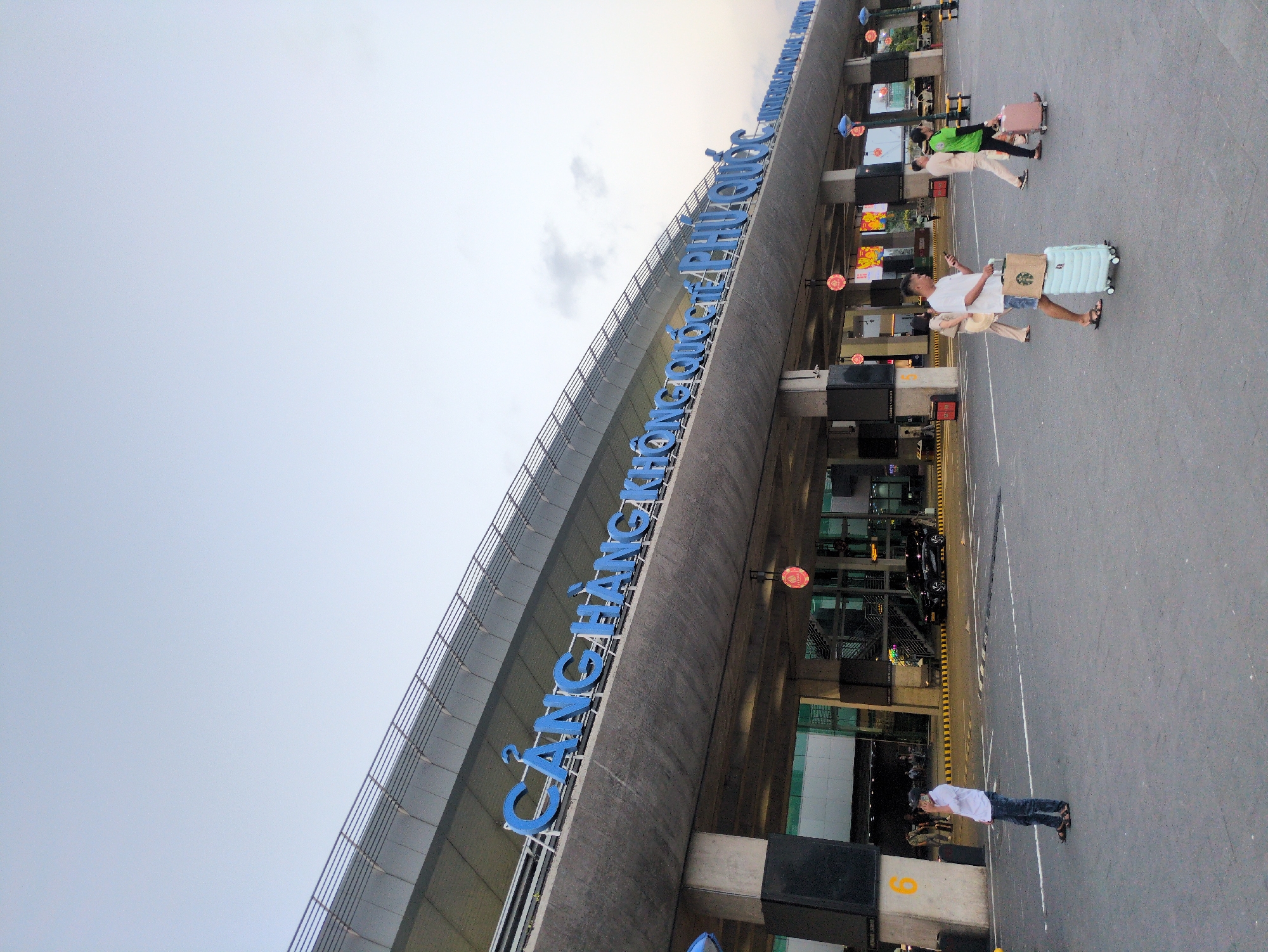 The image depicts the exterior of Cảng Hàng không quốc tế Phú Quốc (Phú Quốc International Airport) in Phú Quốc, Vietnam. People are walking on the pavement in front of the terminal building, some pulling luggage. A man is standing near pillar number 6. The building has blue lettering identifying it. The sky is overcast.