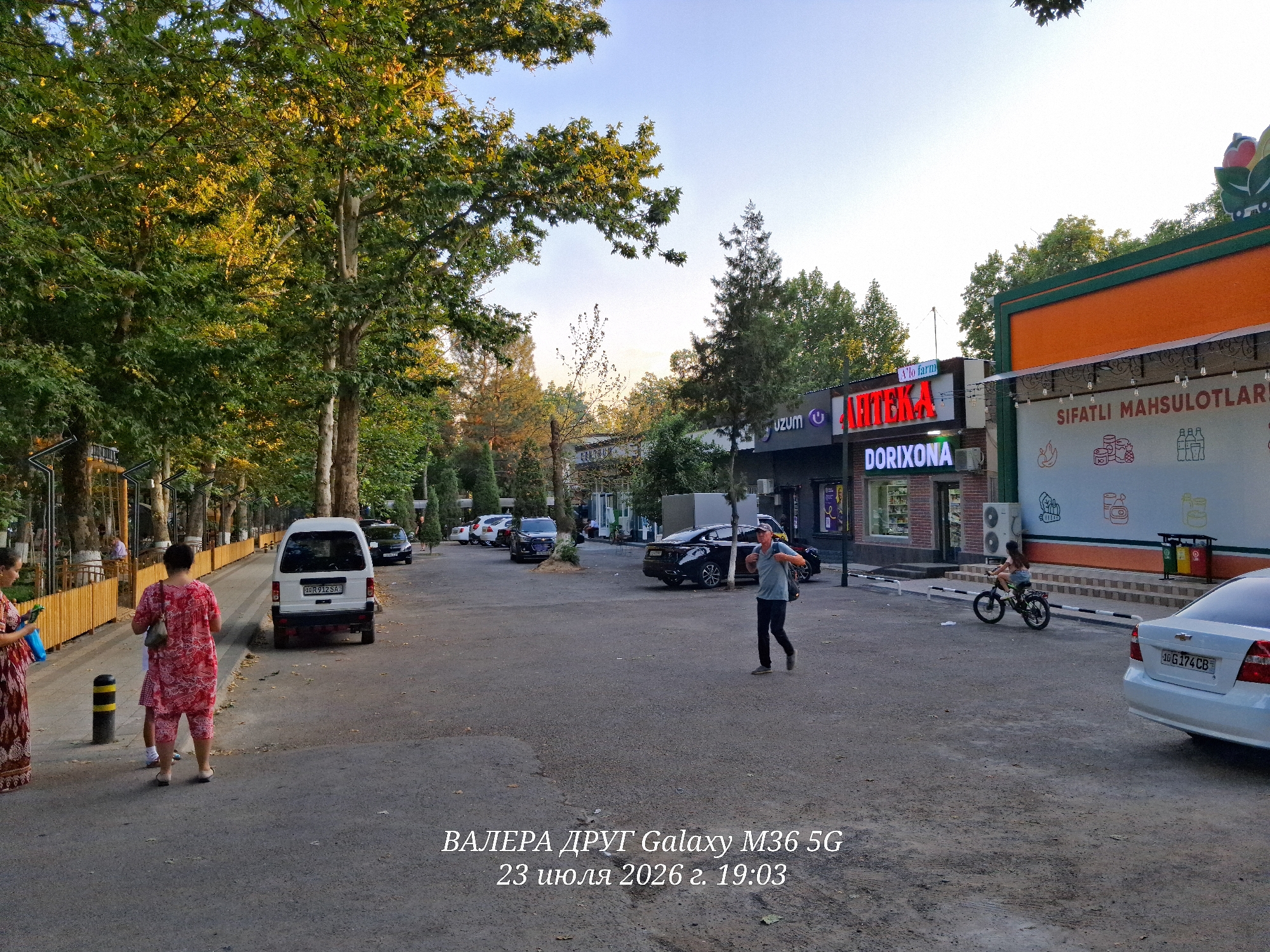 The image depicts a street scene in Chirchik, Uzbekistan. In the foreground, a person is walking with a backpack, while a white car is parked on the right side of the street. Further down the street, several other cars are visible, along with buildings that appear to be shops or businesses, indicated by signage such as "APTEKA DORIXONA" and "SIFATLI MAHSULOTLARI." A young girl is riding a bicycle on the sidewalk. Large trees line the left side of the street, casting shadows on the pavement. The sky is clear and the lighting suggests it is late afternoon or early evening. Text overlay at the bottom of the image indicates "ВАЛЕРА ДРУГ Galaxy M36 5G 23 июля 2026 г. 19:03," possibly a date and time stamp or a caption.