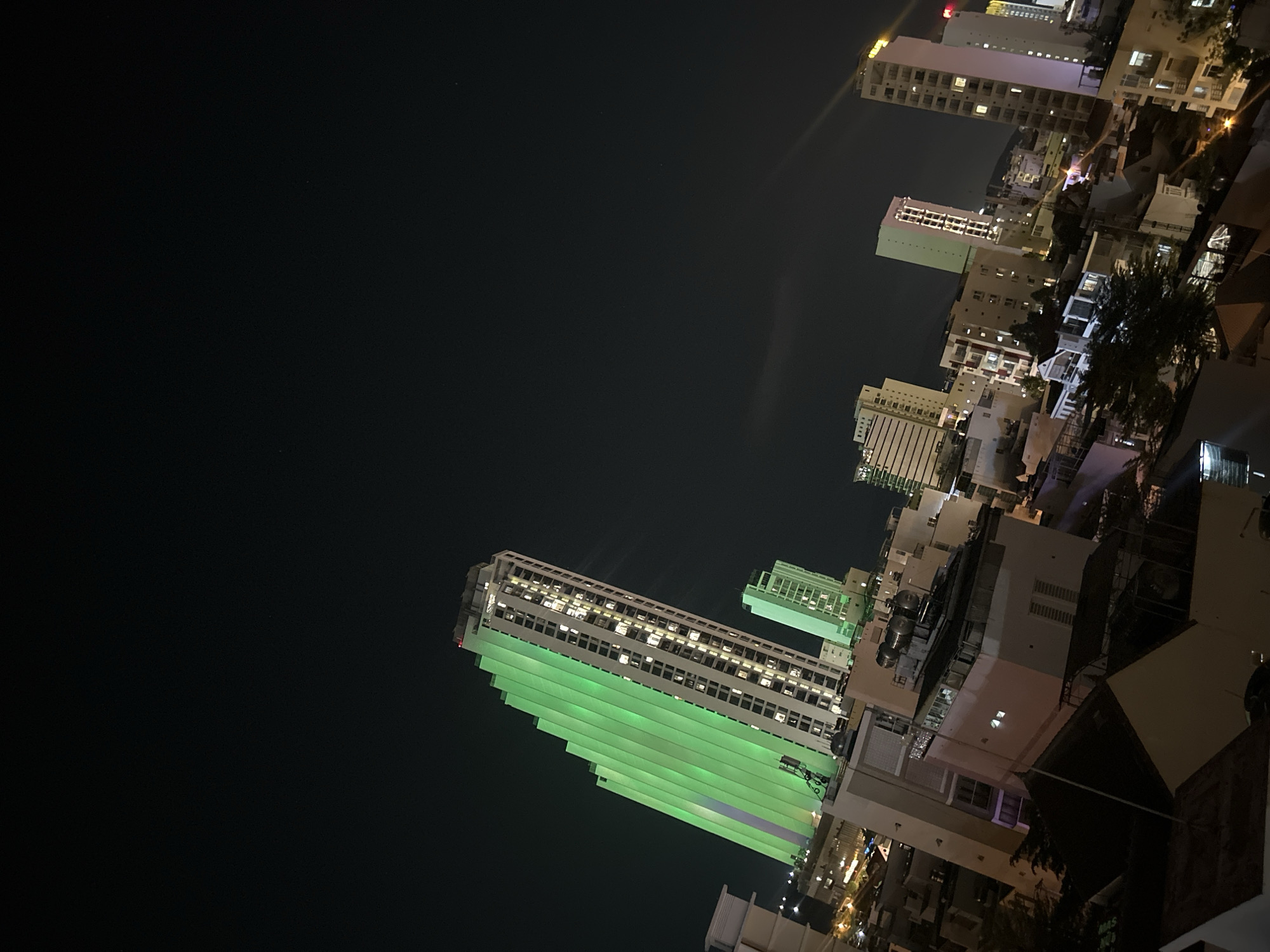 The image shows a nighttime view of Bac Nha Trang Ward, Vietnam. In the lower left, a tall building is lit up with green light on its exterior, with visible windows on the upper section. In the upper right, a cluster of buildings is visible, lit by streetlights and windows. The sky is dark and mostly clear.