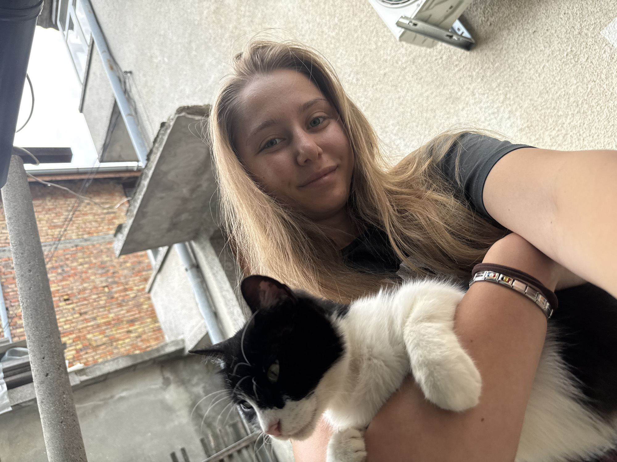 An outdoor, eye-level, close-up selfie shows a young Caucasian woman with long blonde hair and a black t-shirt holding a black and white cat. The woman is smiling at the camera, and the cat is looking down and to the left. The woman is wearing a brown hair tie and a bracelet with multiple charms on her left wrist. The background is blurred, but it appears to be an urban setting with buildings made of brick and concrete, suggesting the location is in Tsarevo, Bulgaria. There is an air conditioning unit visible on the wall in the upper right of the frame.