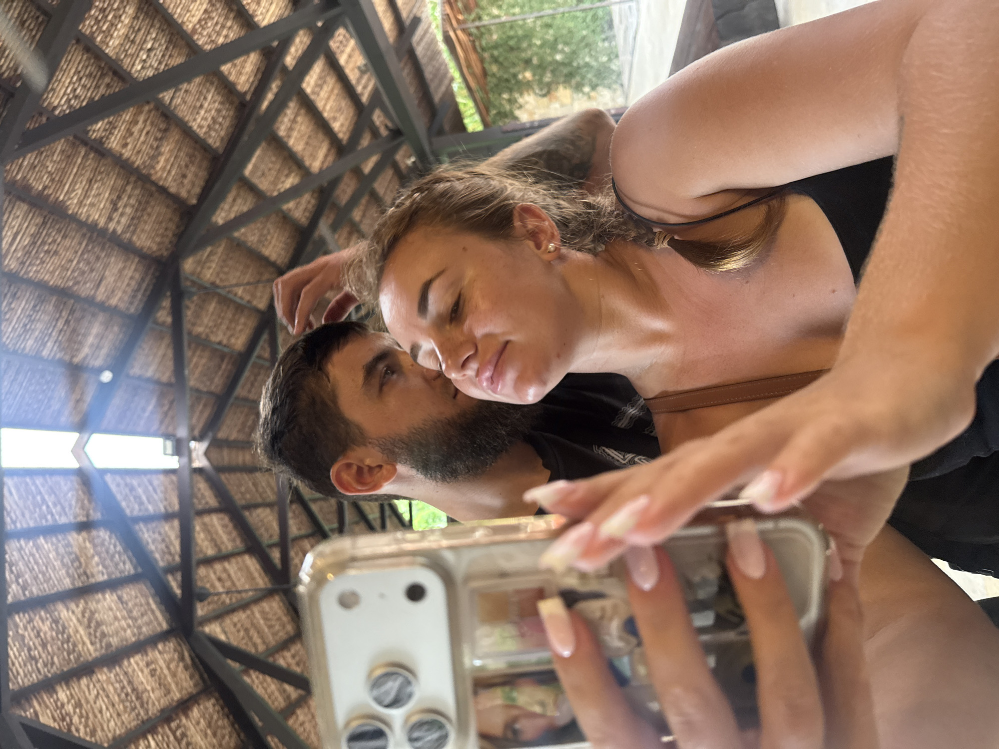 The image shows a couple taking a selfie under a thatched roof structure. The man, with a beard and dark hair, is leaning in close to the woman, touching her hair. The woman, with blonde hair in a braid, is holding a smartphone in her hands, also touching her hair. Both appear to be in a relaxed and intimate pose. The background suggests an outdoor setting, possibly a restaurant or resort, with greenery visible. The architectural style of the roof, with its dark wooden beams and natural thatch, is typical of many coastal resorts and structures in Nha Trang, Vietnam, often found near beaches or tourist areas. The lighting suggests it is daytime, with sunlight filtering through the roof.