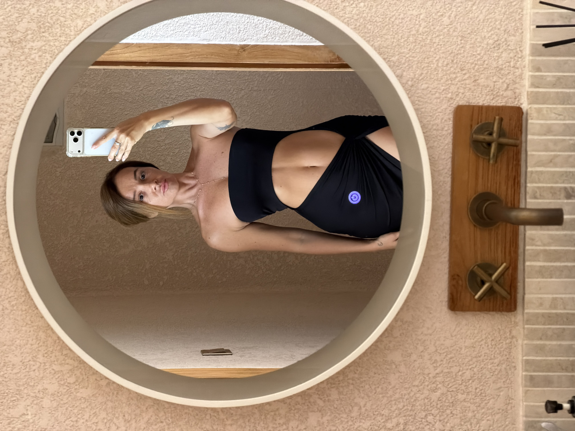 A woman takes a mirror selfie in a bathroom. She is wearing a strapless black dress with a cut-out at the abdomen and a high slit. She has her hair pulled back and is wearing a necklace. The bathroom has a textured pink wall, a round mirror with a light, and wooden accents around the faucets.
