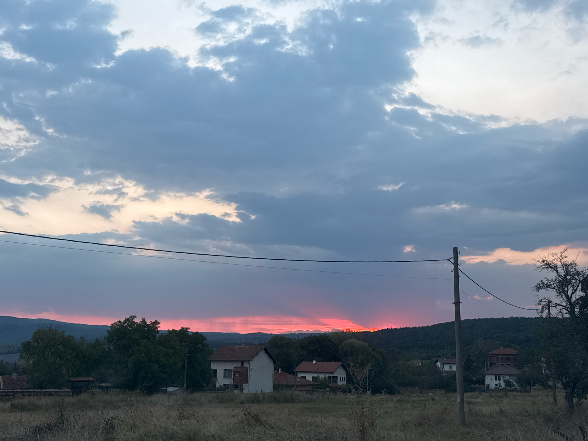 This photograph captures a rural landscape near the city of Verinsko, Bulgaria, during a sunset. The sky is a dramatic canvas of blues, grays, and pinks, with thick clouds obscuring the sun but allowing its vibrant hues to bleed through the lower atmosphere. In the foreground, dry, tall grass covers the ground, suggesting a rural or undeveloped area. Scattered throughout the middle ground are several small houses with red-tiled roofs, typical of Bulgarian villages. Dense green trees dot the landscape between the houses and in the background, forming a dark silhouette against the colorful sky. A utility pole with wires stands prominently on the right side of the image, a common sight in rural areas. The overall scene evokes a sense of quietude and the approaching dusk in a Bulgarian countryside.