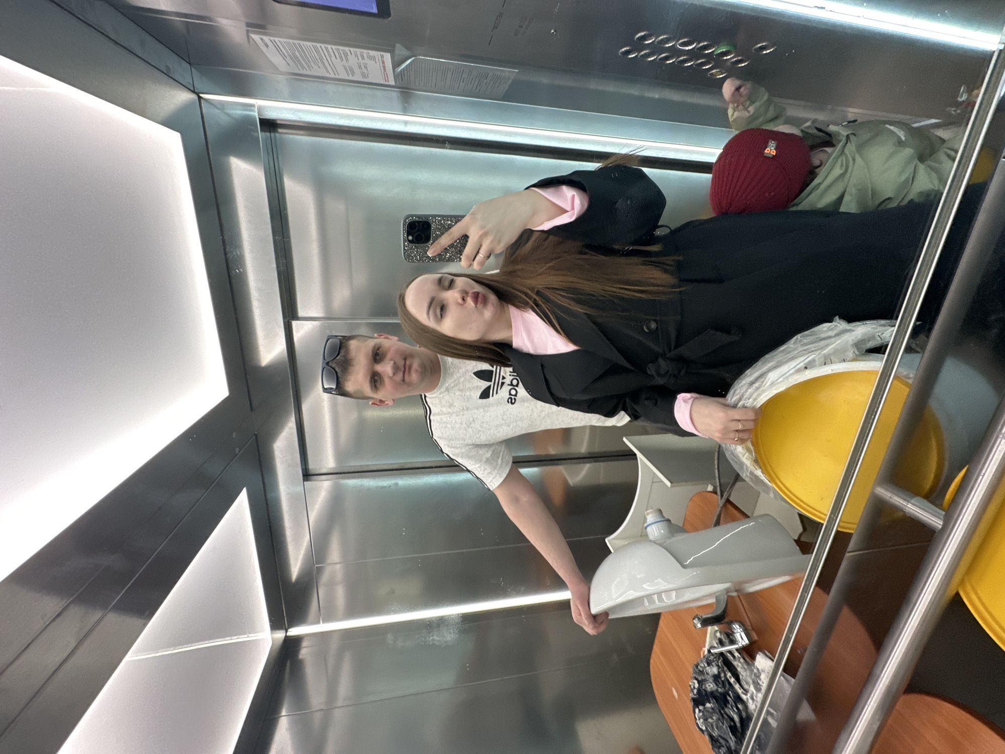 The image depicts three individuals inside a polished metal elevator in Pervomaysky, Russia, transporting household items. In the foreground, a woman with long brown hair, wearing a black trench coat over a pink shirt, is taking a mirror selfie with a smartphone, her lips pursed. Behind and to her left, a man wearing a white t-shirt with an Adidas logo and sunglasses on his head stands, looking towards the camera. His left hand rests on a white porcelain sink that is placed on a light brown wooden surface or table. Next to the sink is a chrome faucet and a crumpled dark fabric. Partially visible in the lower right, a child wearing a red beanie and a green jacket is positioned near the elevator control panel. Two yellow plastic buckets, one wrapped in clear plastic, are visible in the lower portion of the image, below a reflective metal handrail. The elevator features vertical light strips and illuminated ceiling panels.