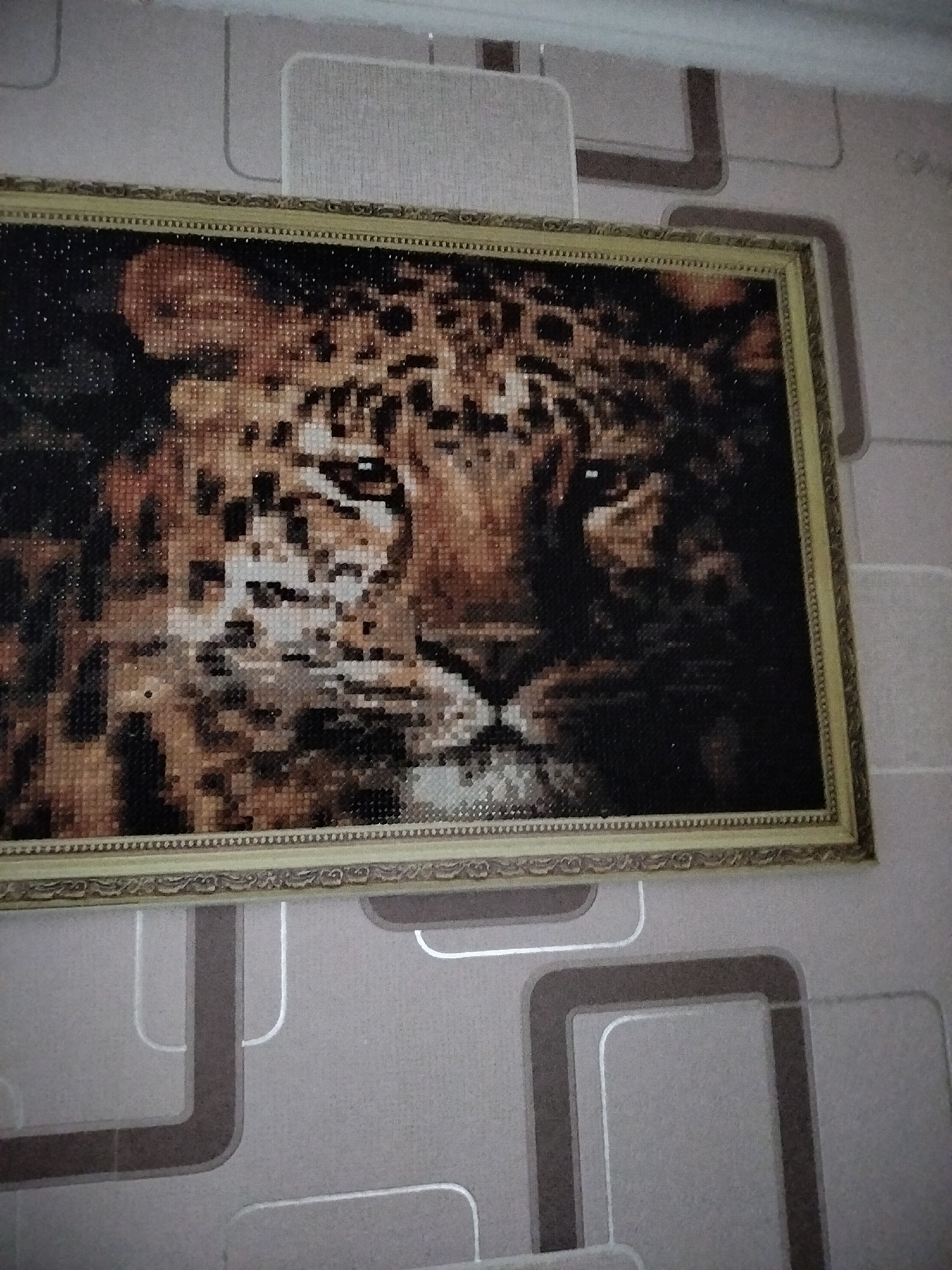 A framed diamond painting of a leopard's face hangs on a wall with grey and beige geometric wallpaper. The painting is rectangular and features a close-up of the leopard's head, with its right side in shadow and its left eye visible. The leopard's fur is rendered in various shades of brown, black, and white, with small, sparkling diamond-like beads. The frame is ornate and golden, with a decorative border. The wallpaper has a repeating pattern of rounded rectangles in shades of grey and beige, with some lines appearing to be outlined in silver. The lighting suggests an indoor setting, possibly a home, given the wallpaper and the framed artwork. The image does not provide information about the city of Nizhnedonskoy, Russia.